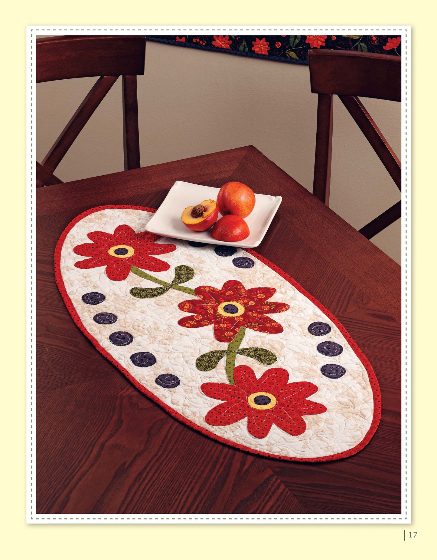 The "Appliqué a Garden" oval table mat by Leisure Arts, featuring red flowers, green stems, and blue circles, sits on a wooden table with a white square plate of three peaches above it—ideal for appliqué projects.