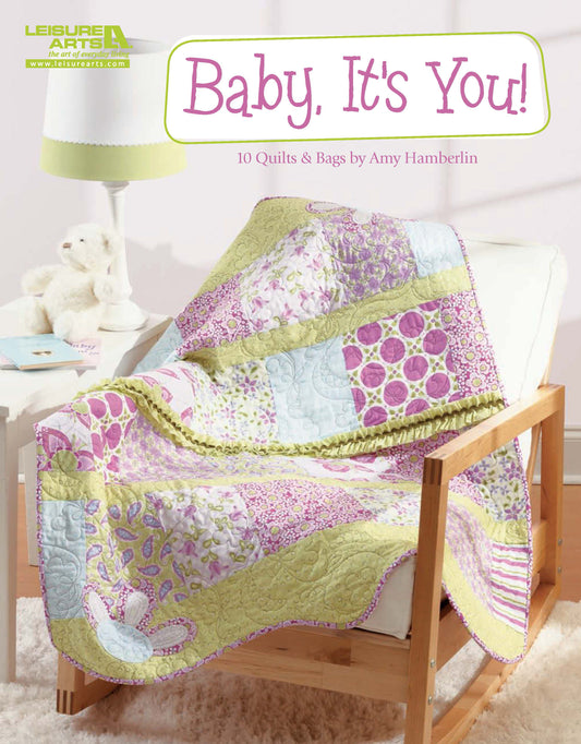 A pastel patchwork quilt with pink, green, and purple patterns drapes over a white chair in a softly lit nursery. On the shelf are decor items and a plush toy. Text: Baby, It's You!—10 Quilts & Bags by Amy Hamberlin (Leisure Arts).