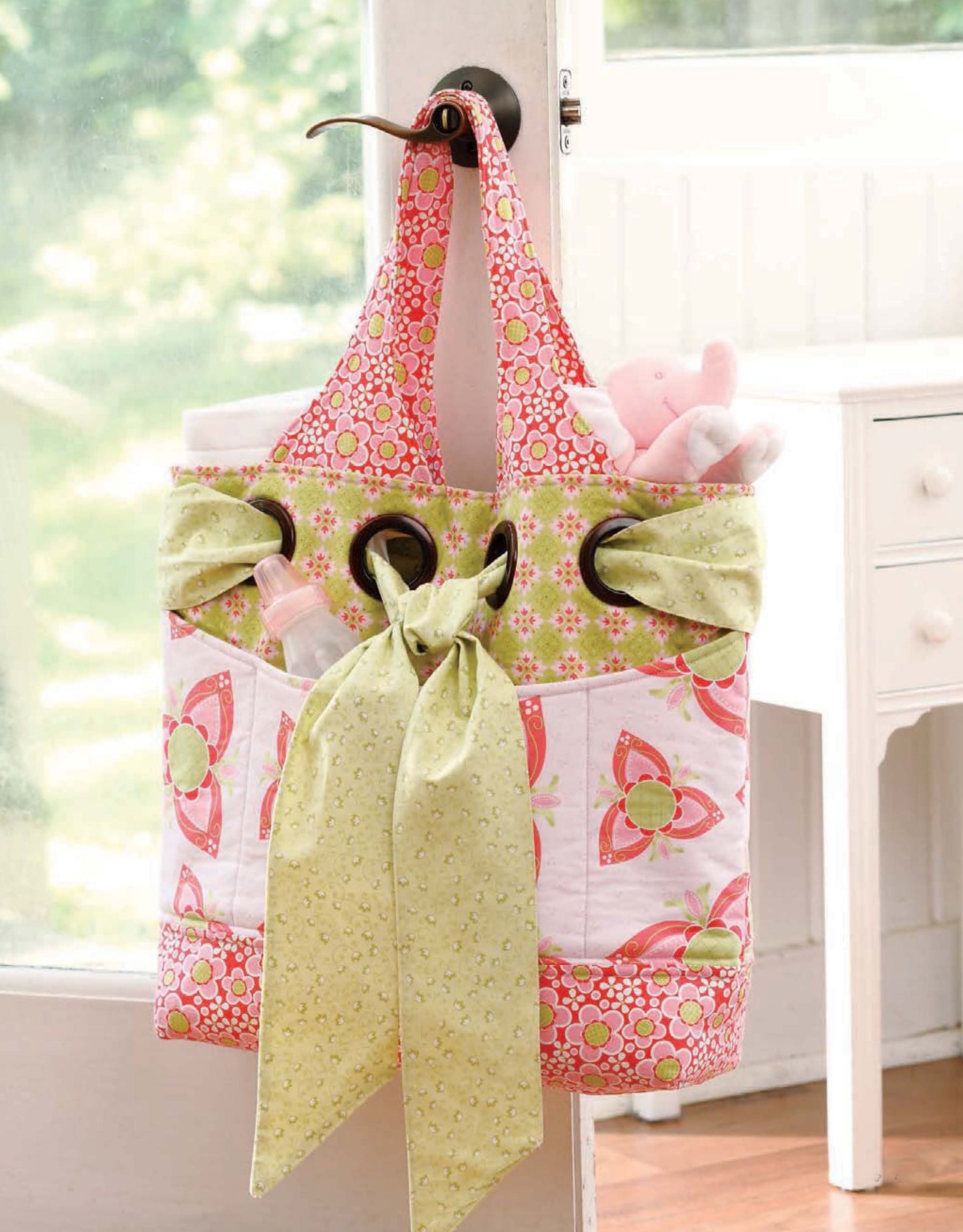 Inspired by baby quilt patterns, the tote bag from "Baby, It's You!—10 Quilts & Bags by Amy Hamberlin" (Leisure Arts) features pink, green, and white floral designs, a large green bow, and holds a stuffed animal, bottle, and blanket.