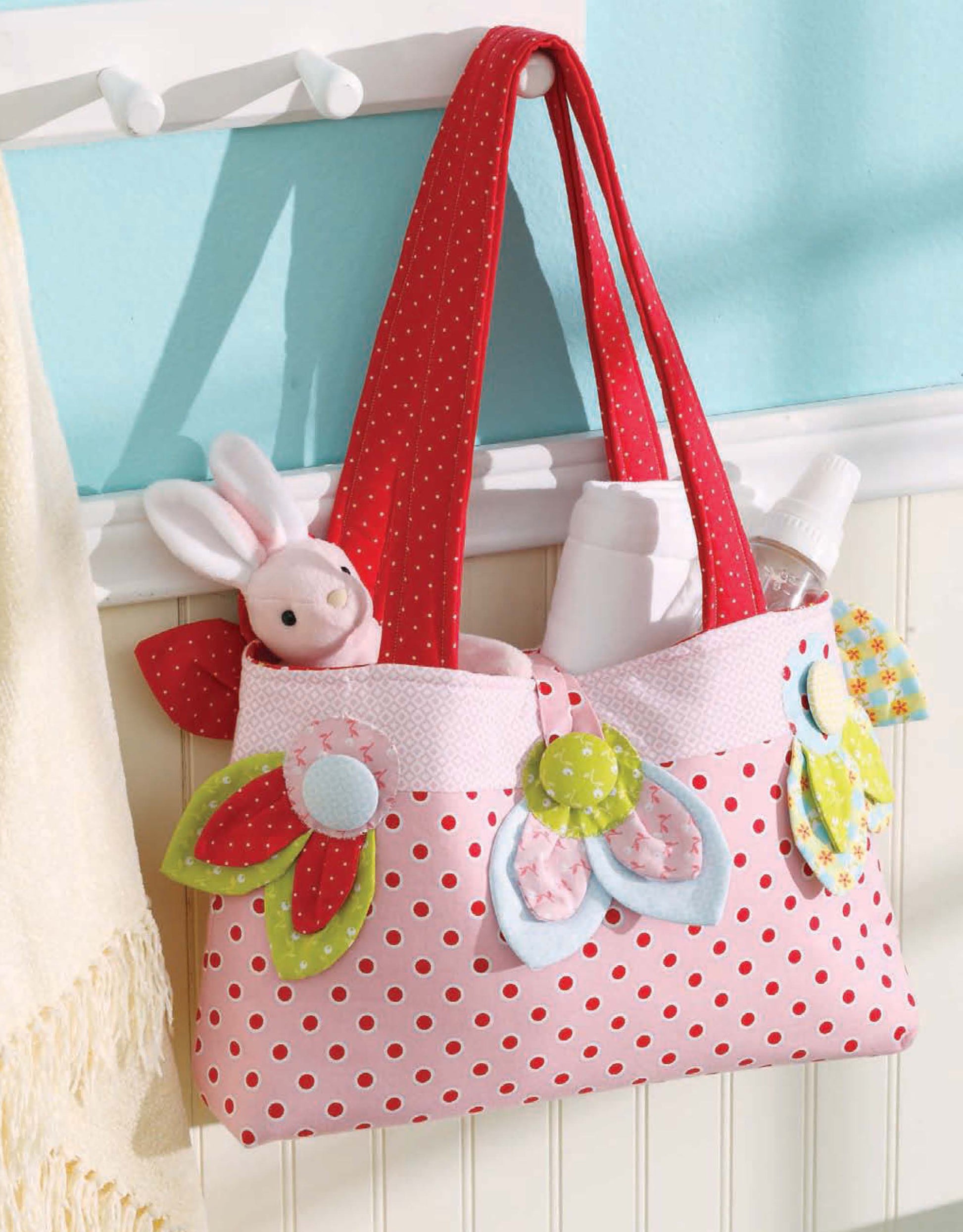 A tote bag from "Baby, It's You!—10 Quilts & Bags by Amy Hamberlin" (Leisure Arts) with polka dots, red handles, and appliqué flowers hangs on a hook, filled with a stuffed bunny, baby bottles, and other baby essentials.