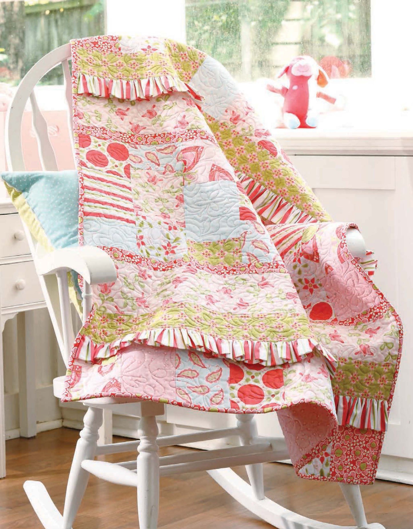 A quilt made with patterns from "Baby, It's You!—10 Quilts & Bags by Amy Hamberlin" (Leisure Arts) drapes over a white wooden rocking chair with a blue pillow. A pink stuffed animal sits on the windowsill in the background.