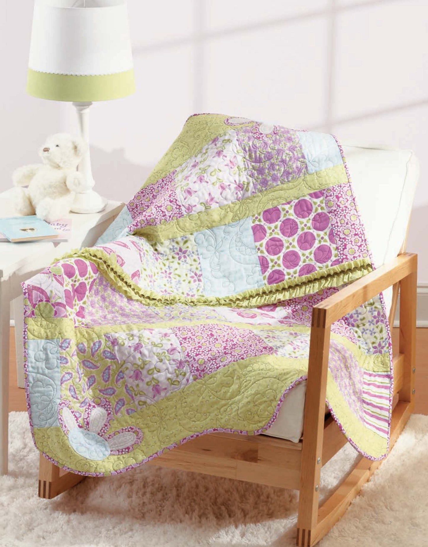 A vibrant quilt from "Baby, It's You!—10 Quilts & Bags by Amy Hamberlin" (Leisure Arts) with pink, purple, green, and blue patterns is draped on a wooden armchair beside a white lamp, plush teddy bear, and shaggy rug in a cozy room.