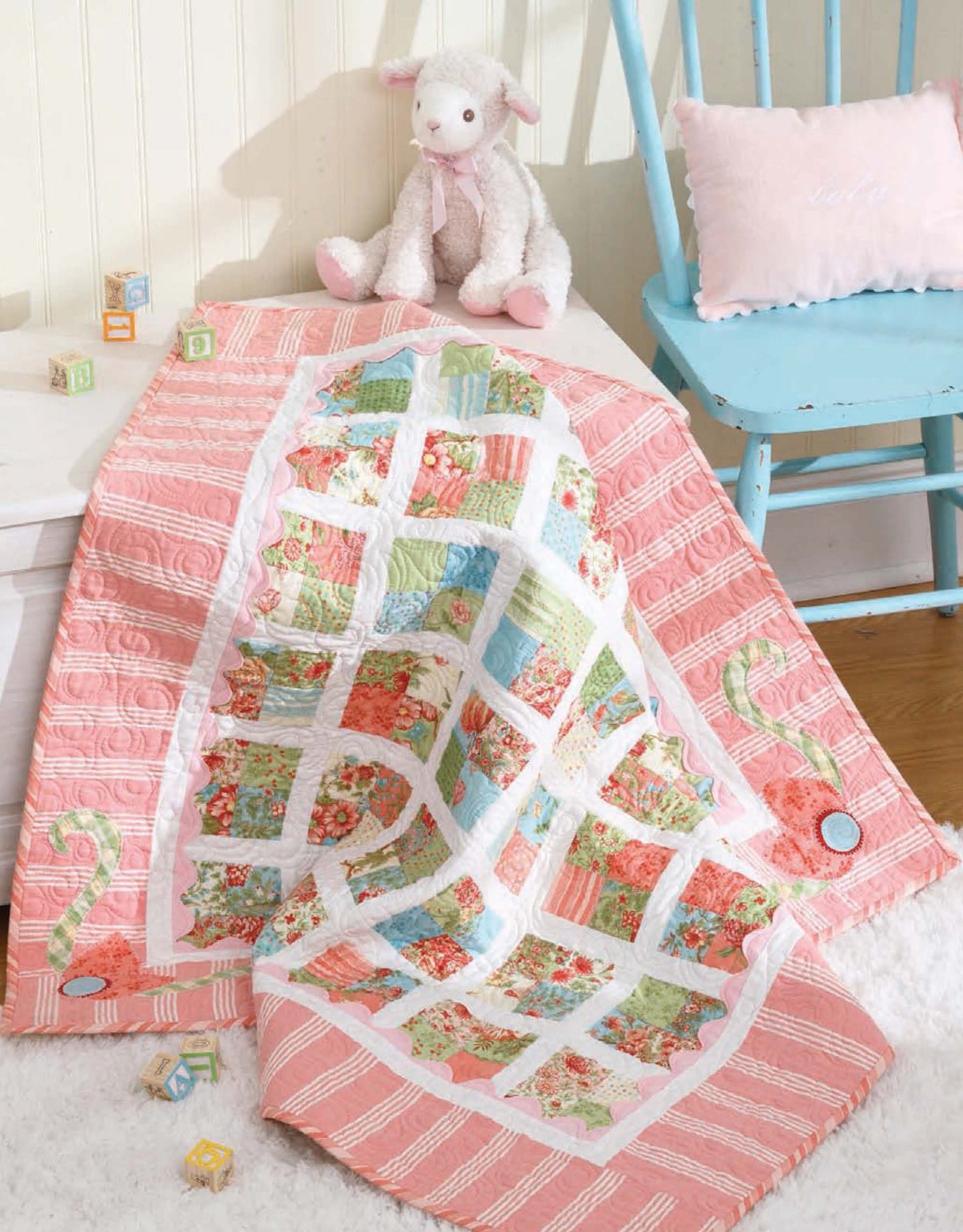 A pastel quilt from "Baby, It's You!—10 Quilts & Bags by Amy Hamberlin" (Leisure Arts) with pink, green, and floral baby patterns is draped over a chair with a pink pillow, beside a white plush lamb and wooden alphabet blocks.