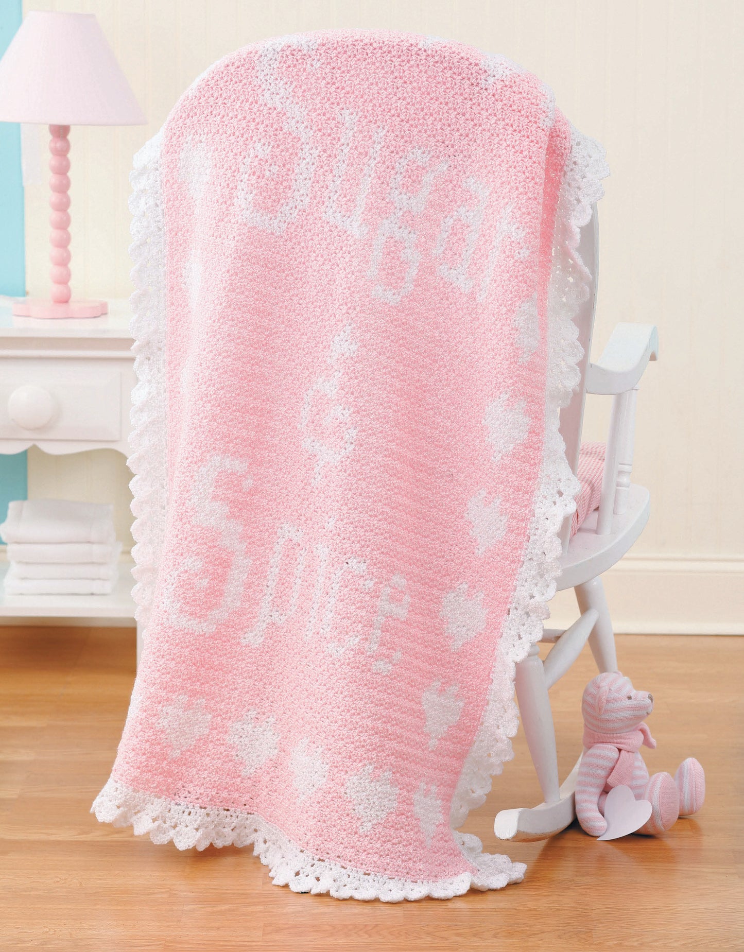 A pink and white crocheted blanket made from Leisure Arts' "A Year of Baby Afghans, Book 5" is draped over a white rocking chair beside a small pink striped stuffed bear on a wooden floor.