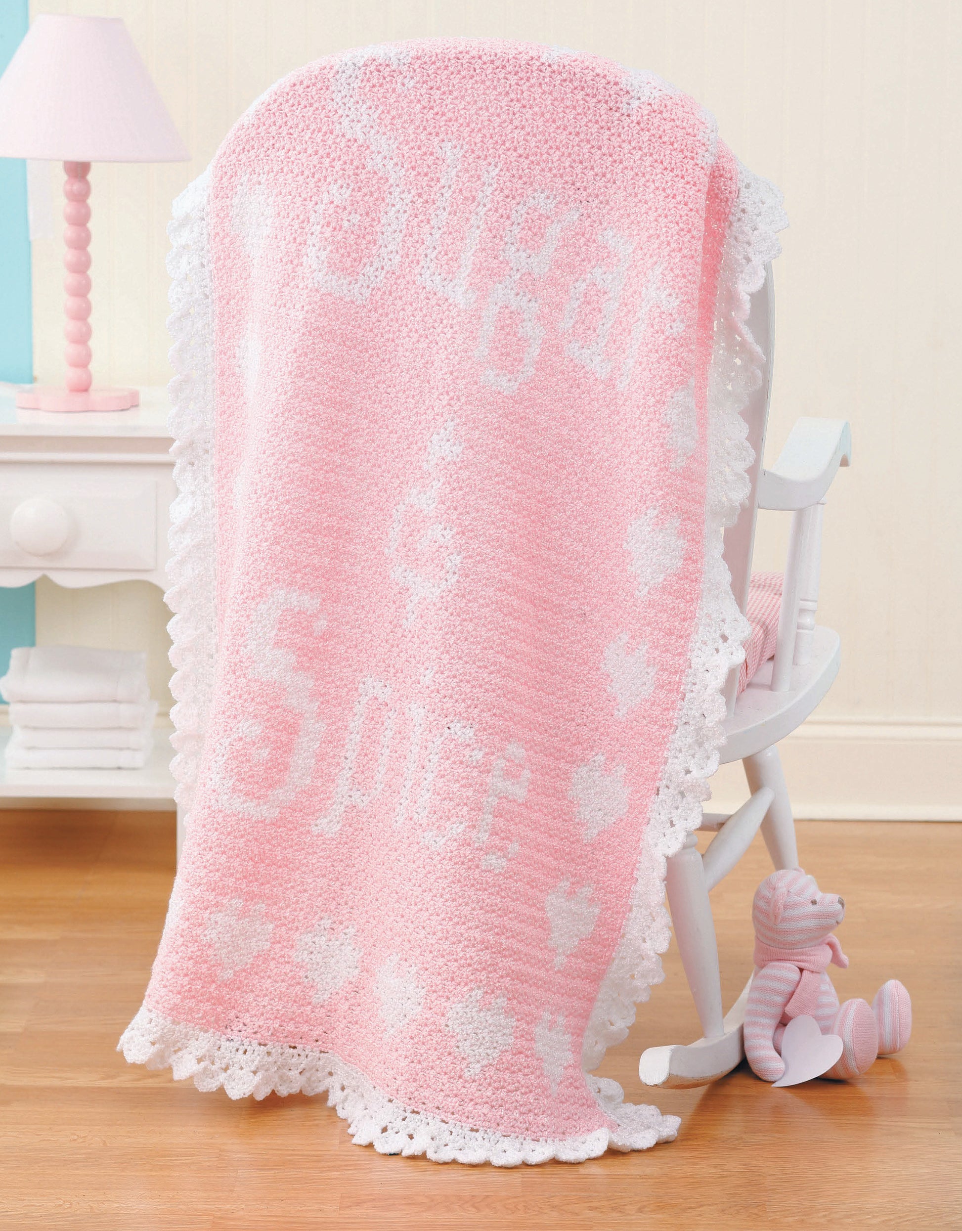 A pink and white crocheted blanket made from Leisure Arts' "A Year of Baby Afghans, Book 5" is draped over a white rocking chair beside a small pink striped stuffed bear on a wooden floor.