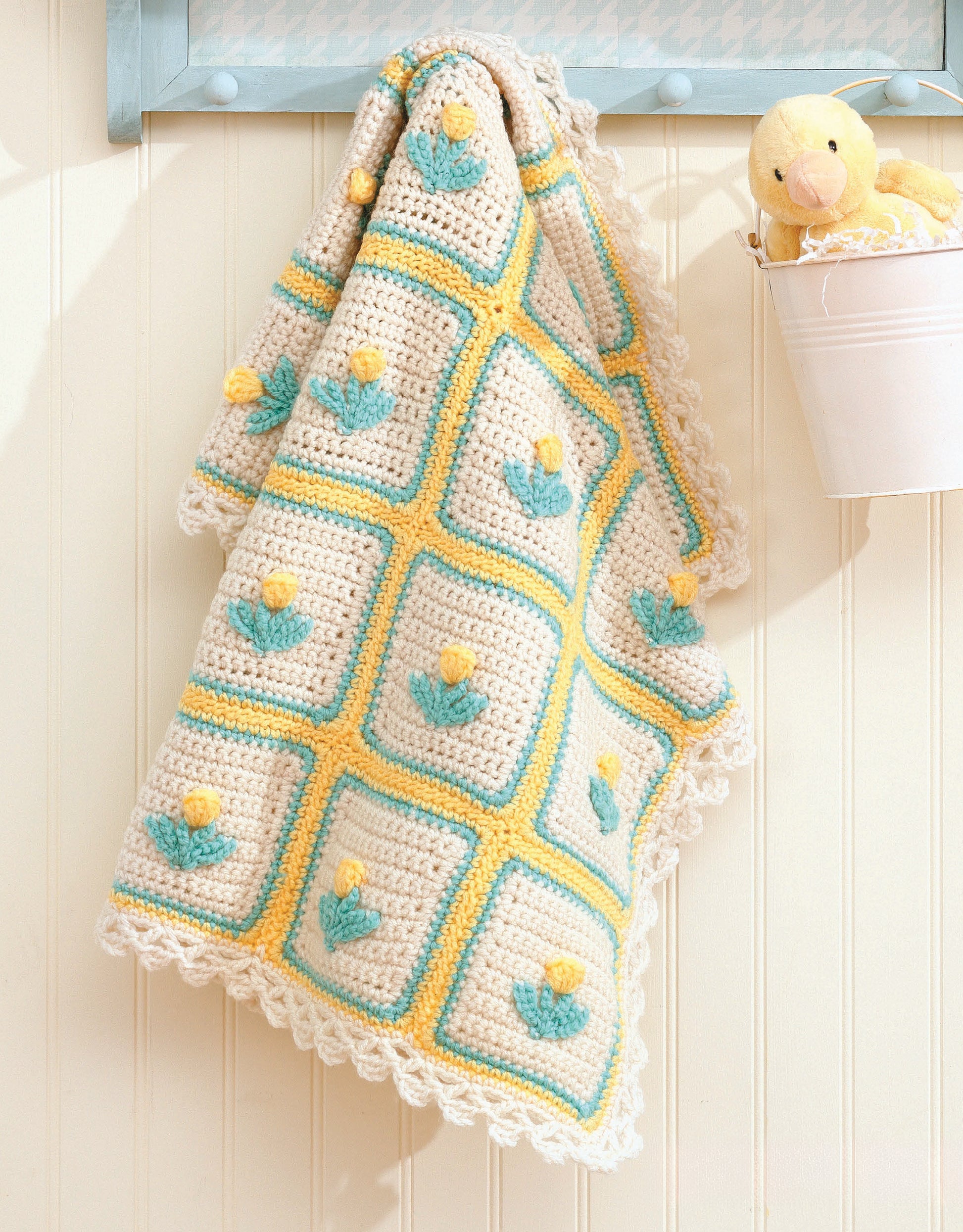 A crocheted baby blanket made from "A Year of Baby Afghans, Book 5" by Leisure Arts, with yellow, blue, and white flowered squares, hangs on the wall. A plush duck toy sits in a white pail nearby for a charming nursery touch.