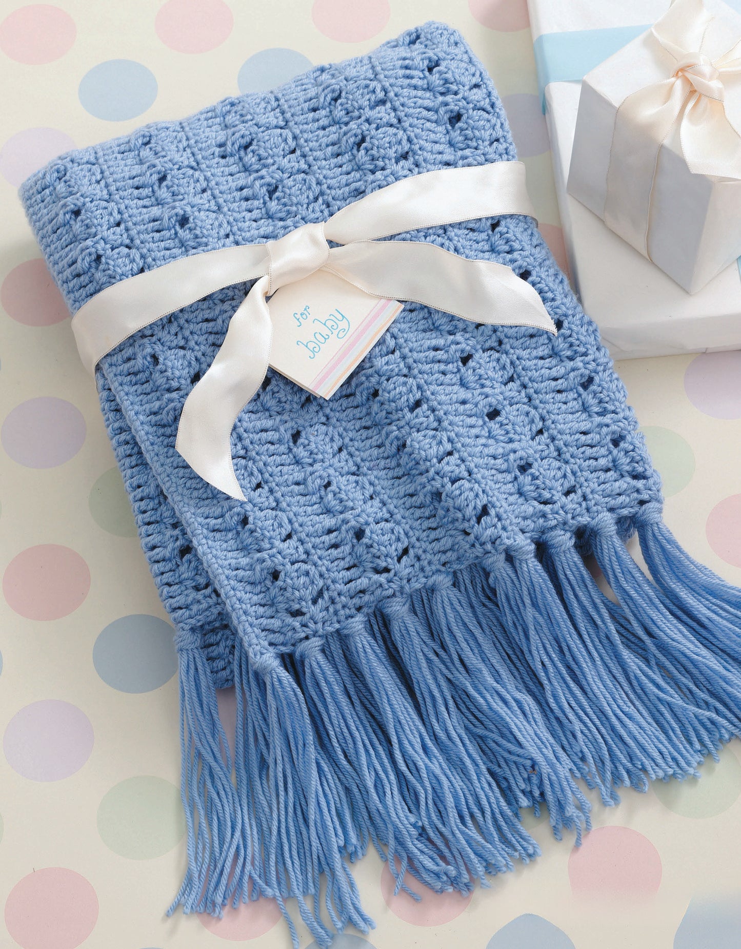 A folded blue crocheted baby blanket with tassels and a “for baby” tag sits beside a white gift box, inspired by patterns in "A Year of Baby Afghans, Book 5" from Leisure Arts—perfect for your next crochet project.