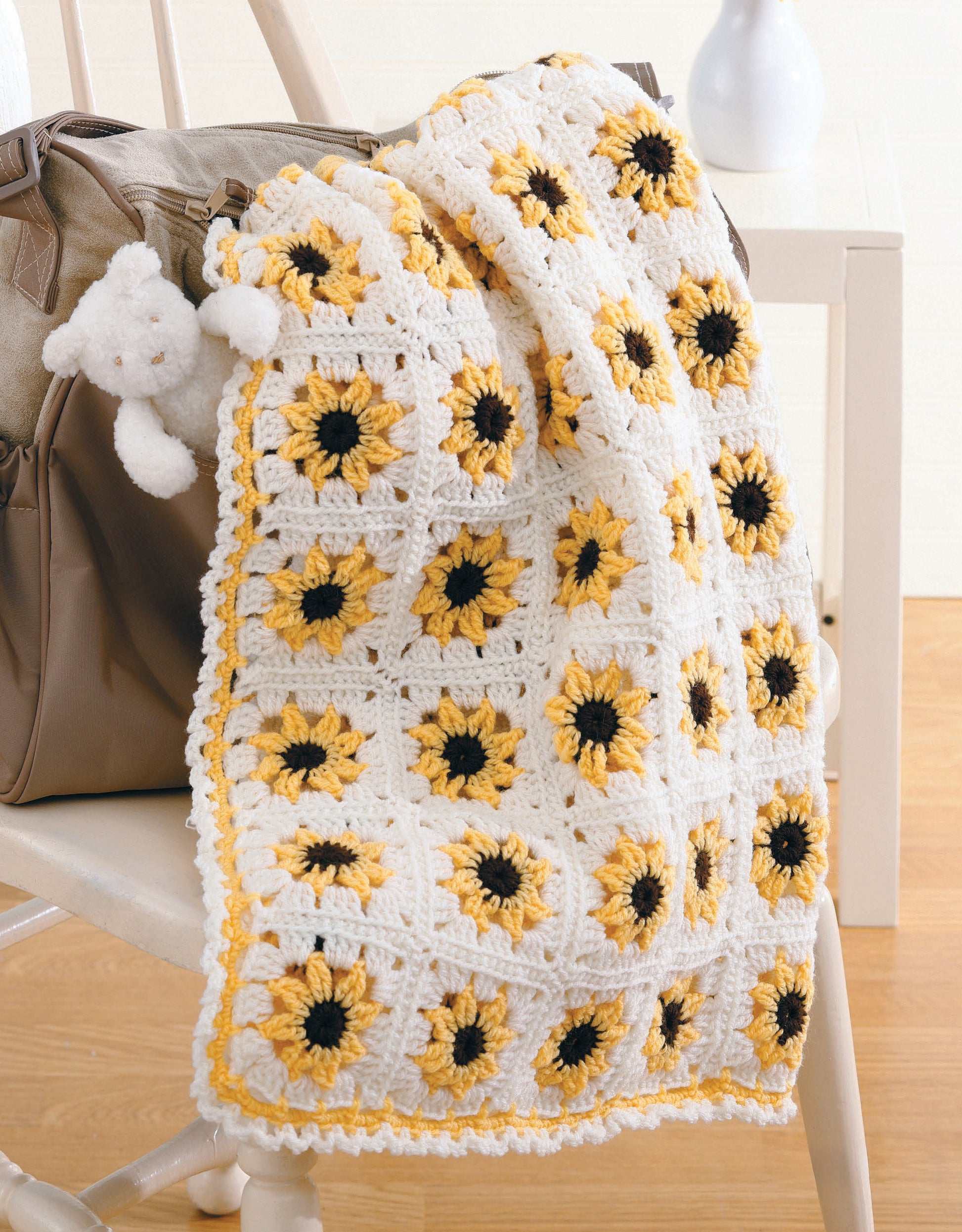 A Year of Baby Afghans, Book 5 by Leisure Arts features colorful crochet designs—a sunflower-patterned blanket in yellow, white, and black draped over a chair next to a beige bag with a small white teddy bear attached.