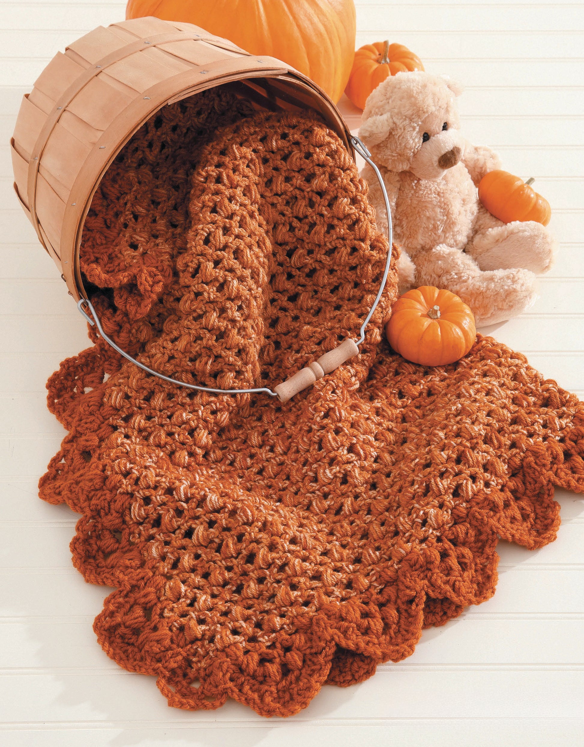 A cozy crocheted blanket from "A Year of Baby Afghans, Book 5" by Leisure Arts spills from a wooden basket, surrounded by small pumpkins and a plush teddy bear on a light surface.