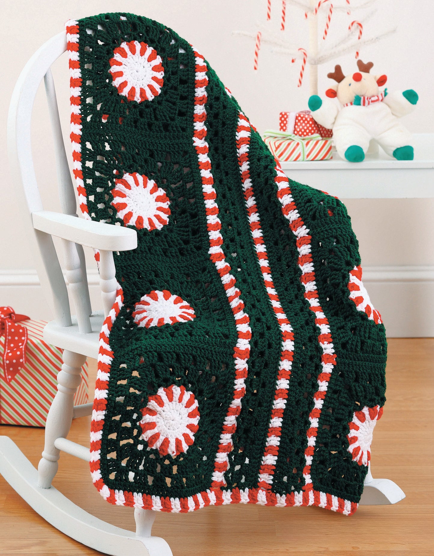 A Year of Baby Afghans, Book 5 by Leisure Arts features a green, white, and red striped crocheted blanket with circles draped over a white rocking chair amid holiday décor—perfect for colorful baby blanket inspiration.