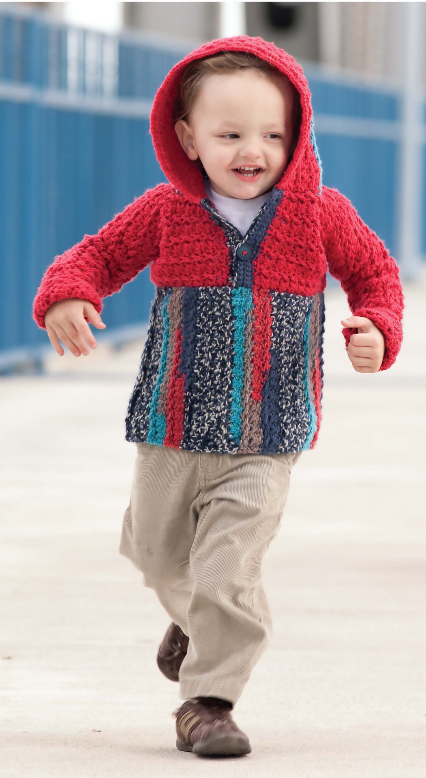A young child, smiling as they run outdoors in a red and multicolored knit hoodie made from worsted weight yarn inspired by Leisure Arts’ Blossom Street Collection Book 1, with beige pants and brown shoes. Blue railings blur in the background.