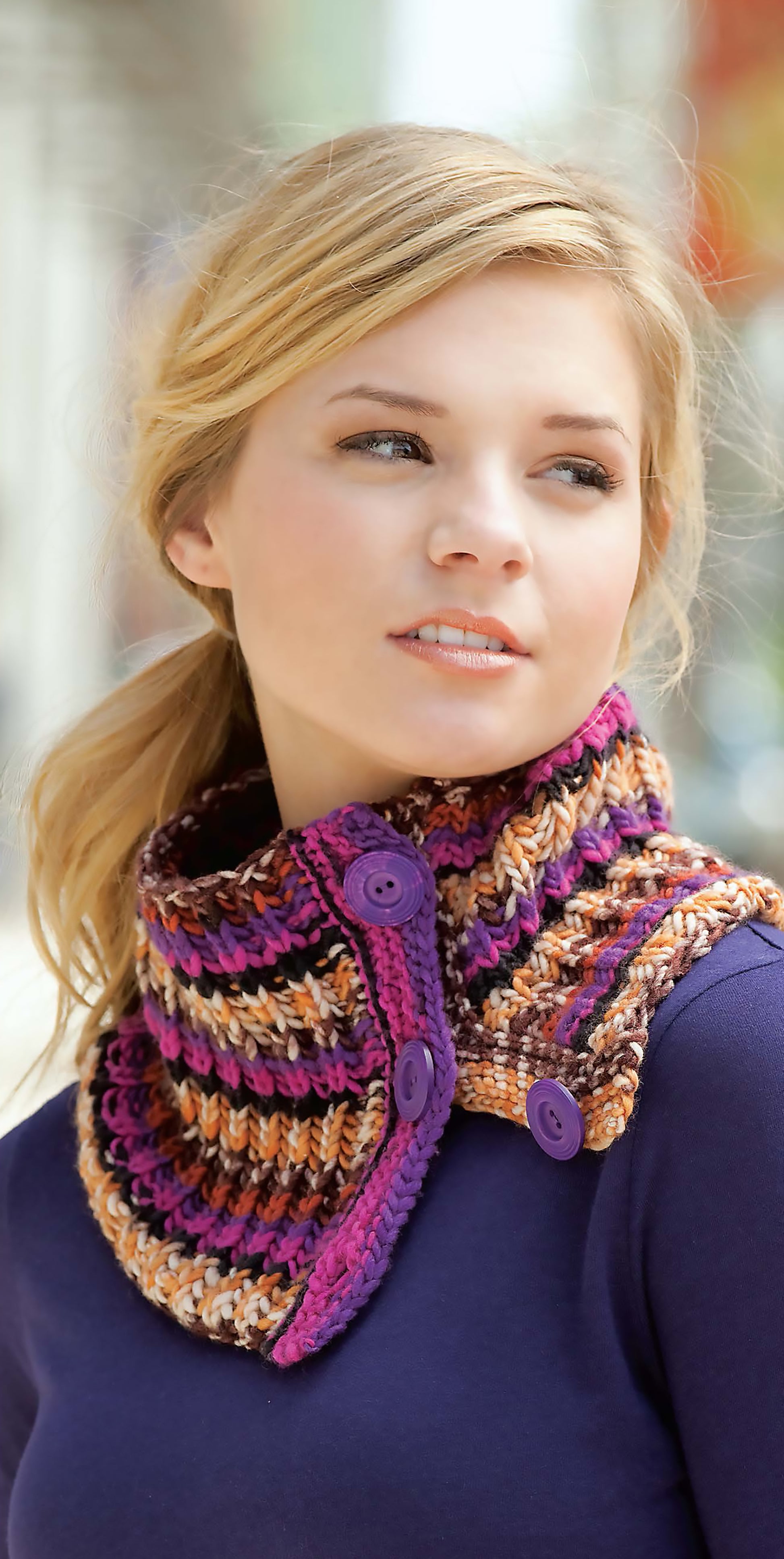 A young woman with blonde hair in a loose ponytail wears a knitted, multicolored neck warmer made from patterns in Blossom Street Collection Book 1 by Leisure Arts, paired with a navy blue top, and looks to the side thoughtfully.
