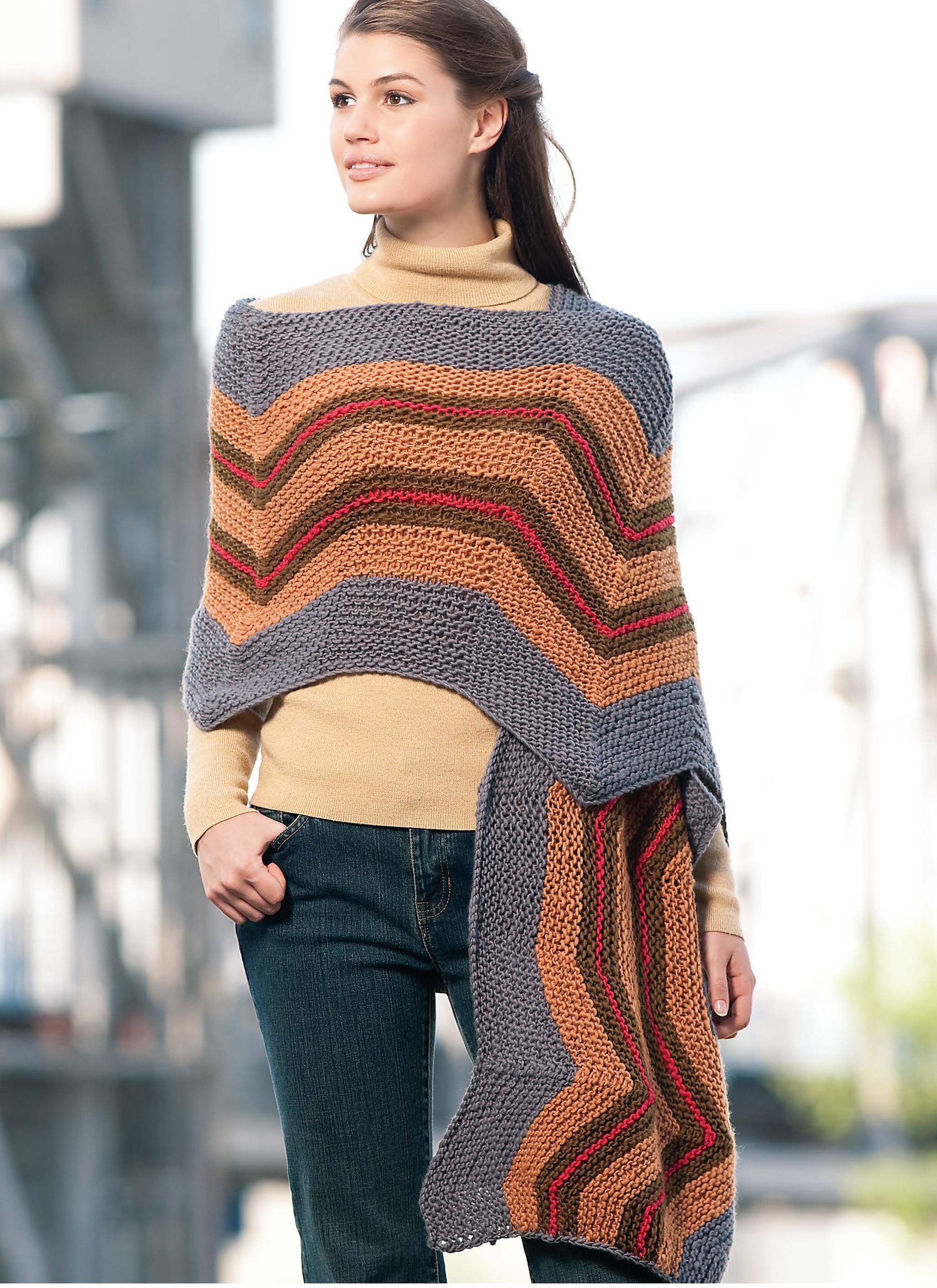 A woman in a tan turtleneck and jeans poses outdoors, draped in a chevron-striped shawl made with worsted yarn. The shawl pattern is from the Blossom Street Collection Book 1 by Leisure Arts; an industrial scene blurs in the background.
