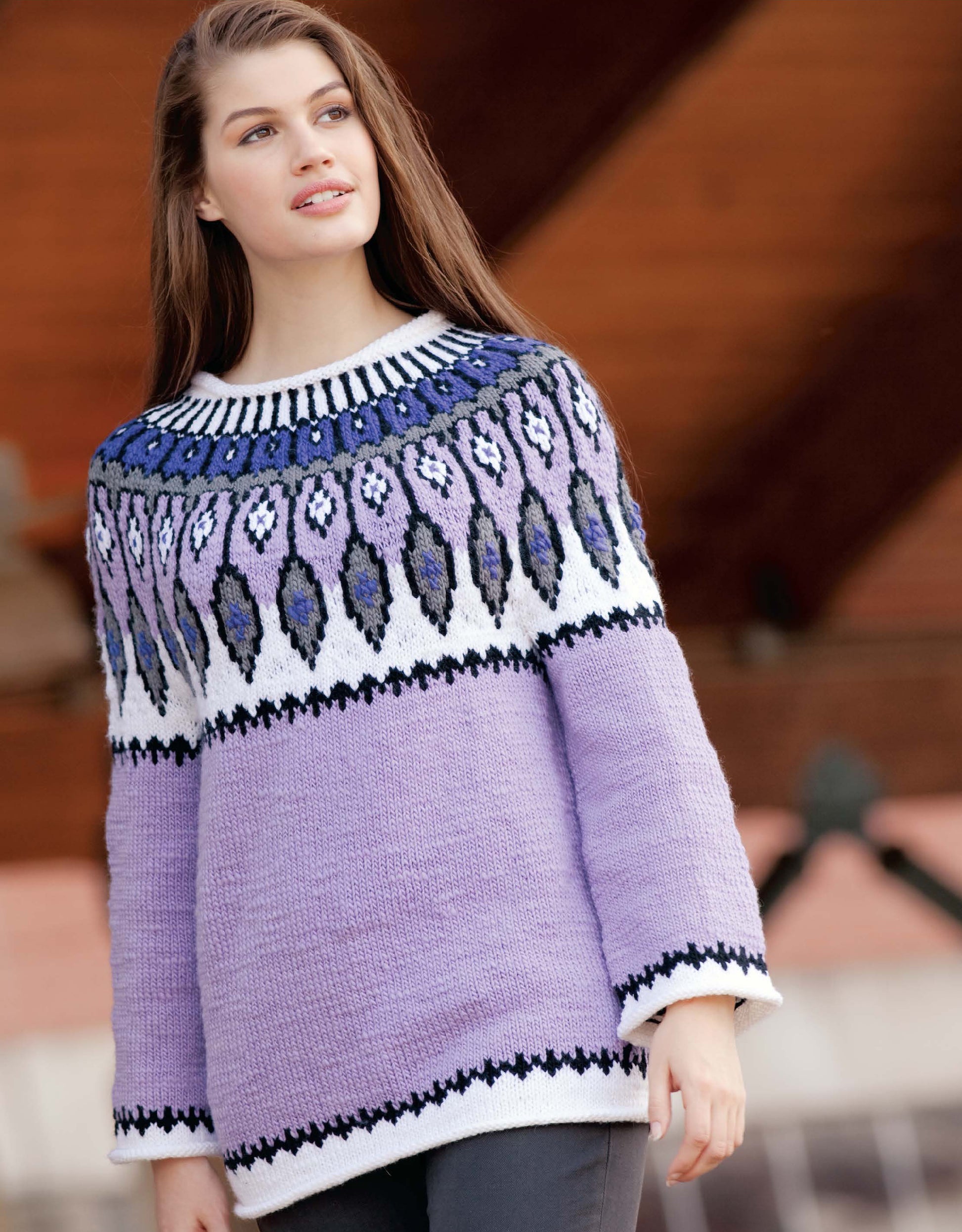 A woman with long brown hair models a lavender worsted weight sweater with a blue, white, and purple yoke from the Blossom Street Collection Book 1 by Leisure Arts, standing outdoors before a blurred wooden backdrop.