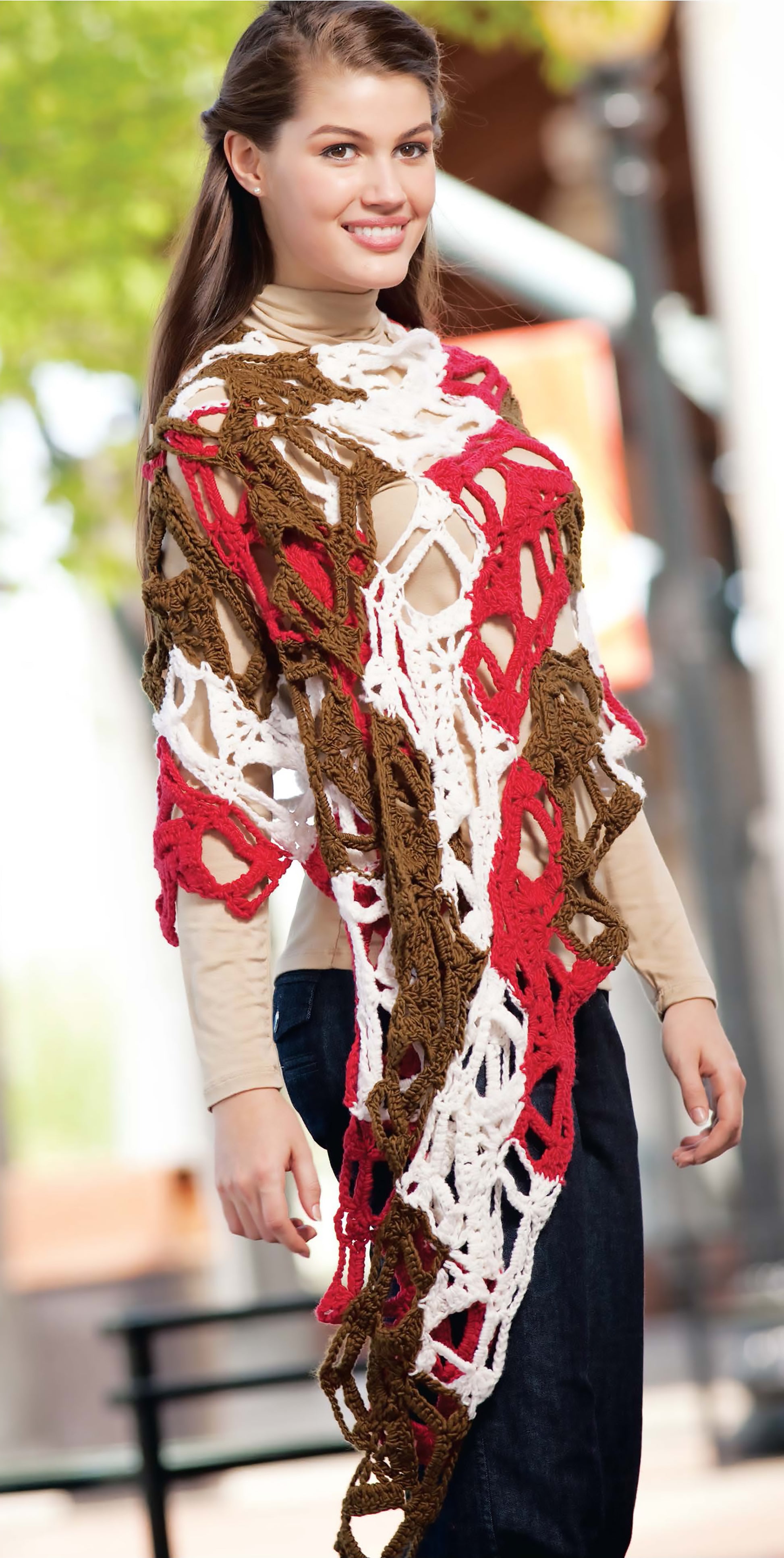 A woman models a loosely crocheted red, white, and brown shawl made with worsted yarn from the Blossom Street Collection Book 1 by Leisure Arts, worn over a beige shirt and jeans against an outdoor backdrop of trees and buildings.