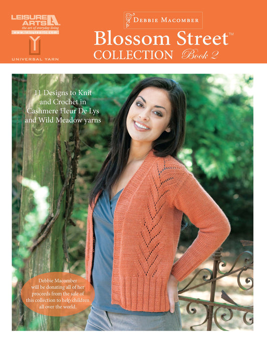 A smiling woman wearing a peach knit cardigan stands by a wooden fence. She holds Leisure Arts’ Blossom Street Collection Book 2 Digital Download, featuring easy knitting projects and step-by-step instructions for knitting and crochet.