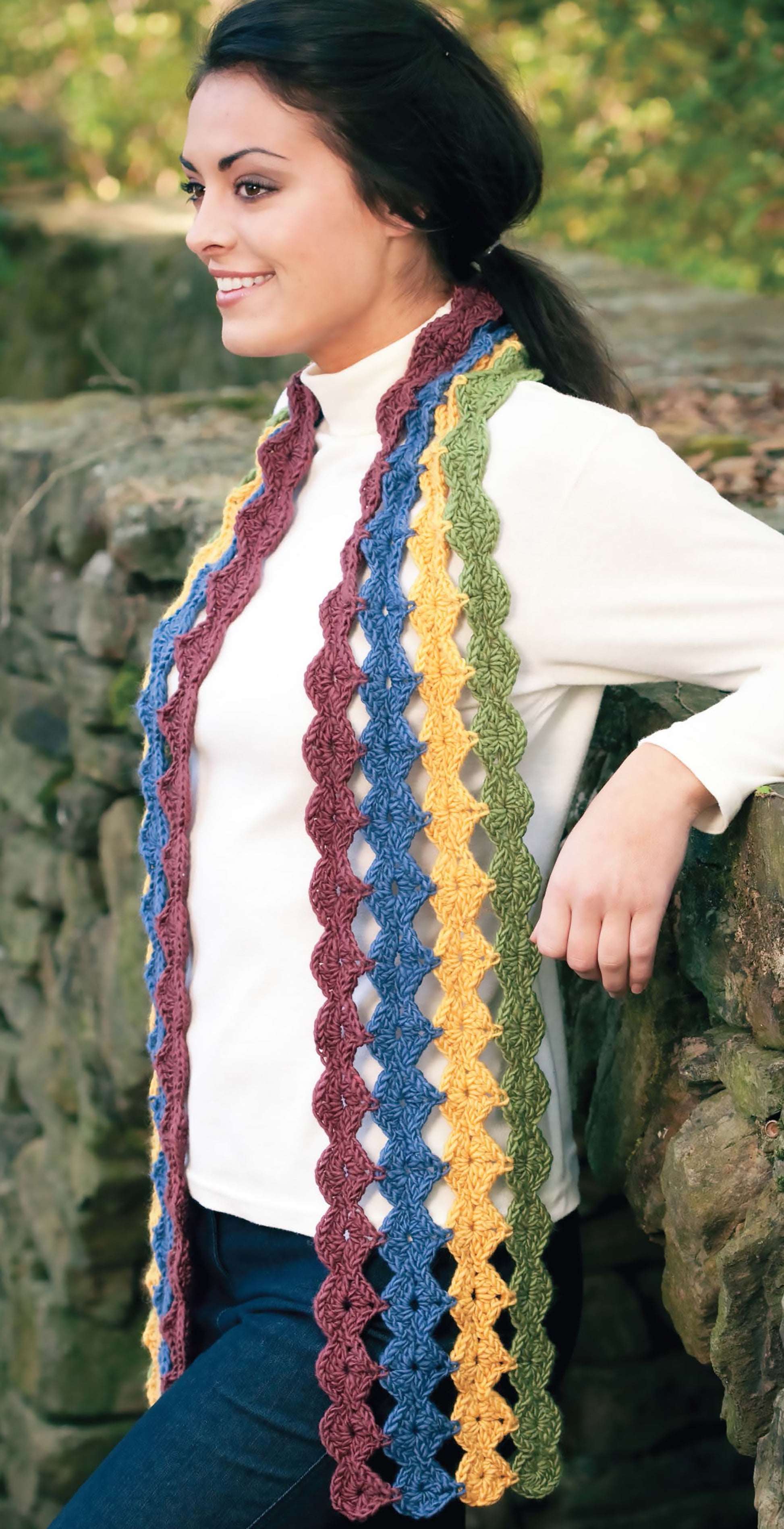 A woman in a white turtleneck smiles outdoors, wearing a vibrant crochet scarf. Get inspired for your next project with the Blossom Street Collection Book 2 Digital Download from Leisure Arts!.