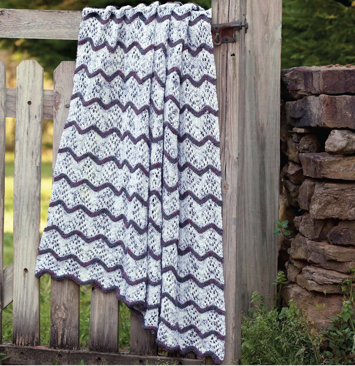 A knitted blanket with a wavy pattern in white, purple, and gray hangs over a rustic gate. Get inspired with easy projects from Blossom Street Collection Book 2 by Leisure Arts, perfect for knitting cozy blankets or cardigans.