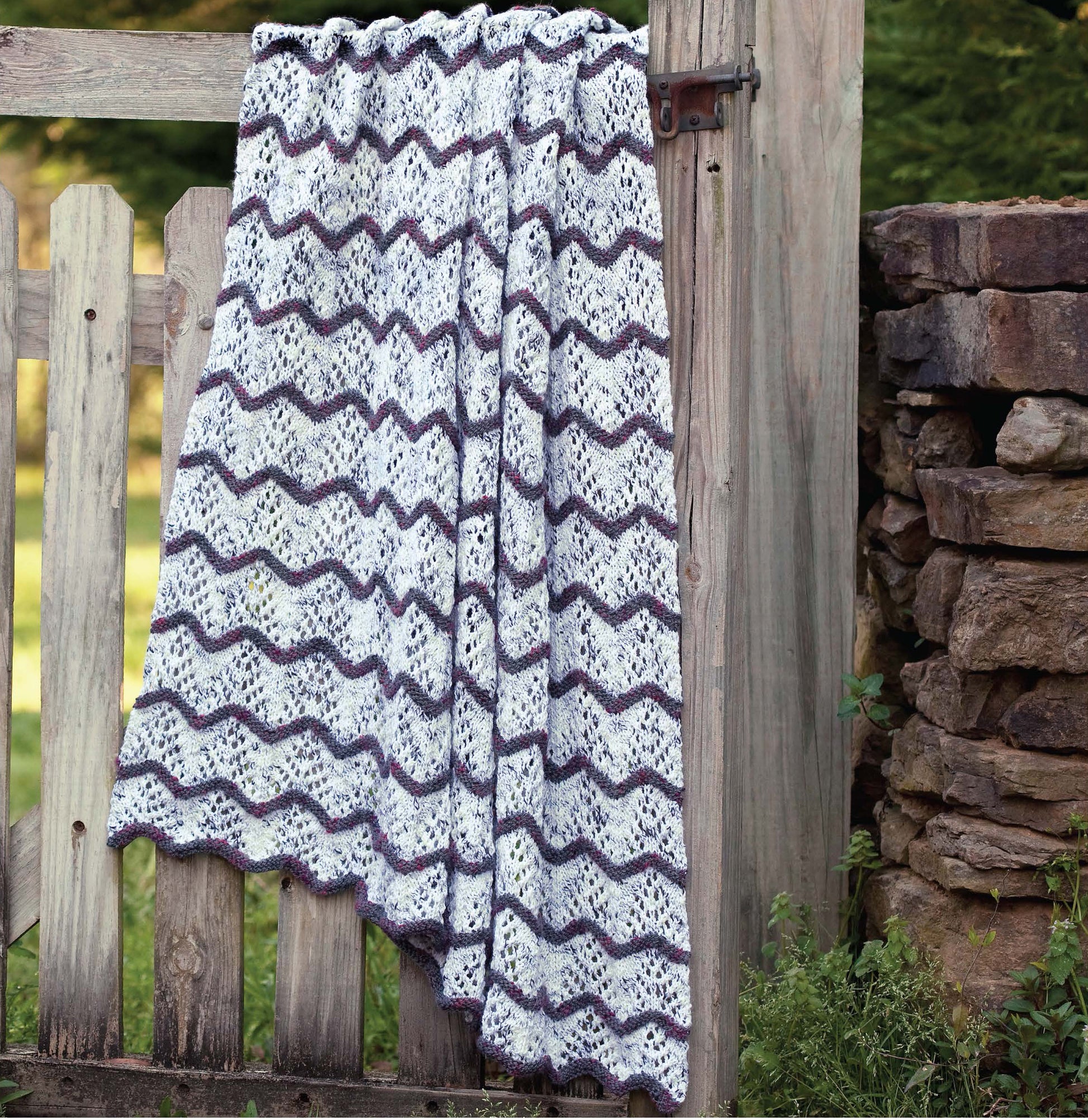 A knitted blanket with a wavy pattern in white, purple, and gray hangs over a rustic gate. Get inspired with easy projects from Blossom Street Collection Book 2 by Leisure Arts, perfect for knitting cozy blankets or cardigans.