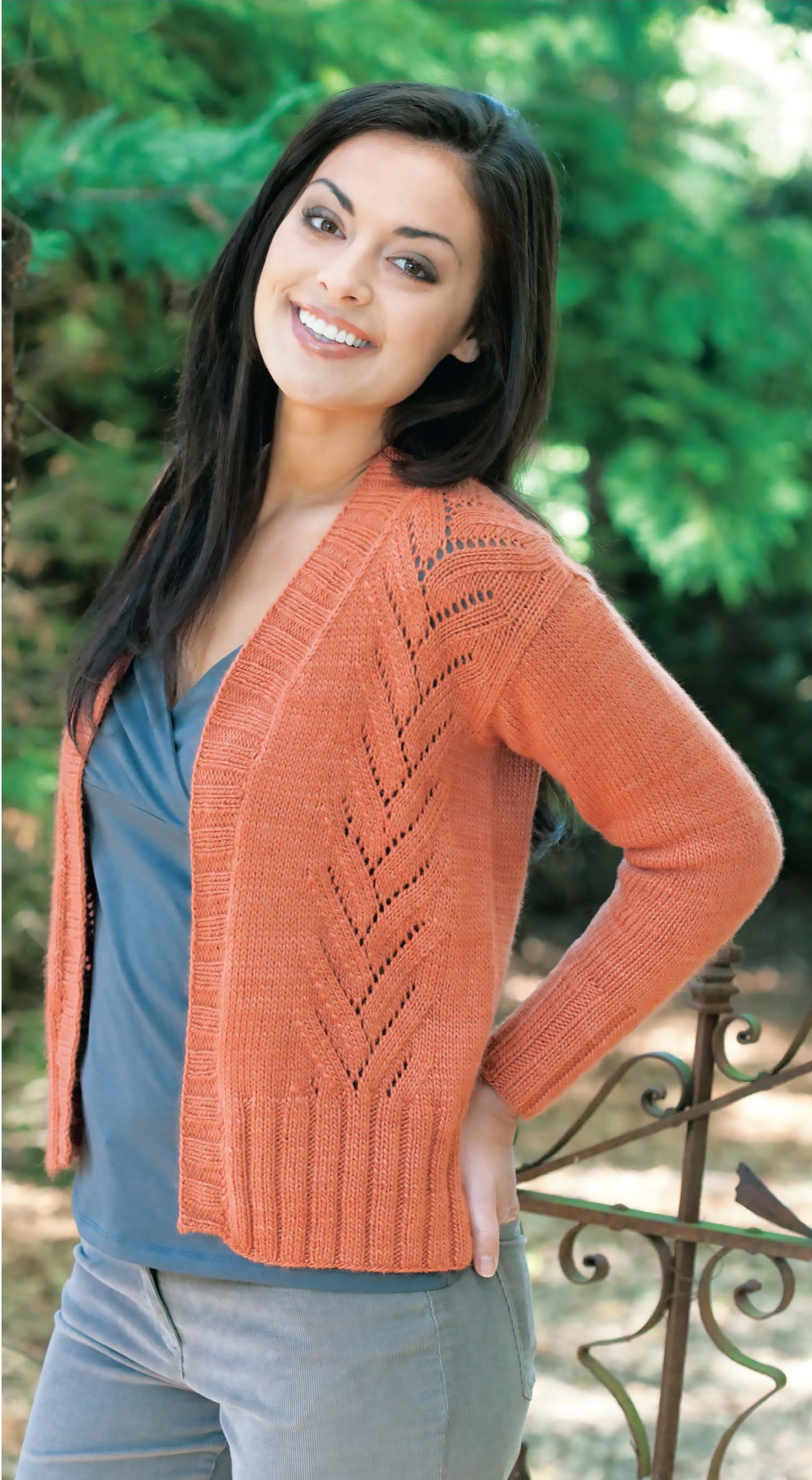 A woman with long dark hair smiles outdoors, wearing a rust-colored, leaf-patterned cardigan—an easy project inspired by the Blossom Street Collection Book 2 Digital Download from Leisure Arts. Lush green trees and a wrought iron fence are behind her.