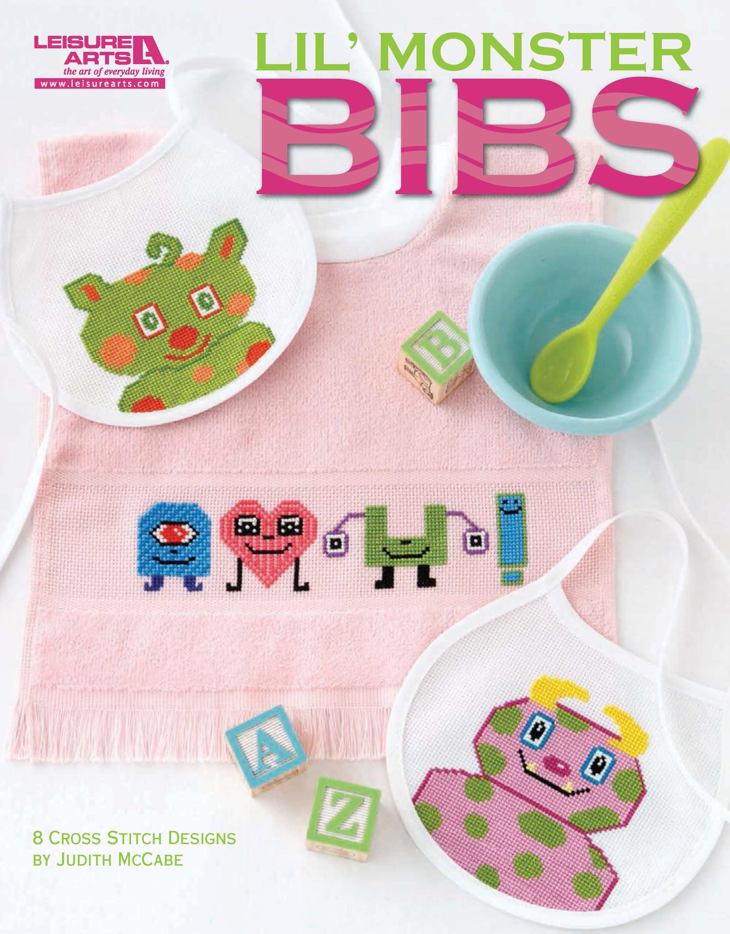 A magazine cover shows three white Aida bibs with colorful monster cross stitch patterns beside a pink baby bowl, spoon, and pastel alphabet blocks. The title "Lil' Monster Bibs" features this Leisure Arts digital download.