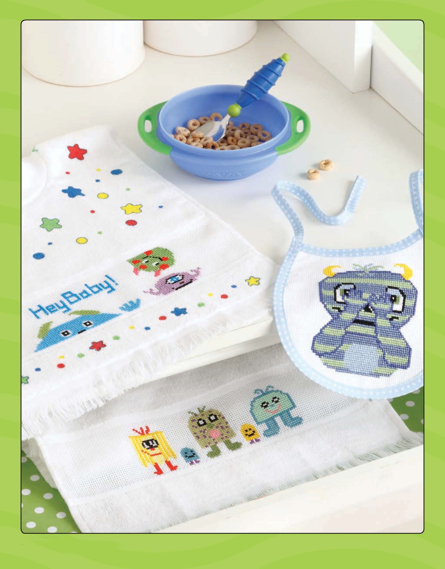 Leisure Arts' Lil' Monster Bibs Digital Download features colorful cartoon monster embroidery, perfect for cross stitch. A blue cereal bowl with a green-handled spoon accents the playful baby set on a white surface.