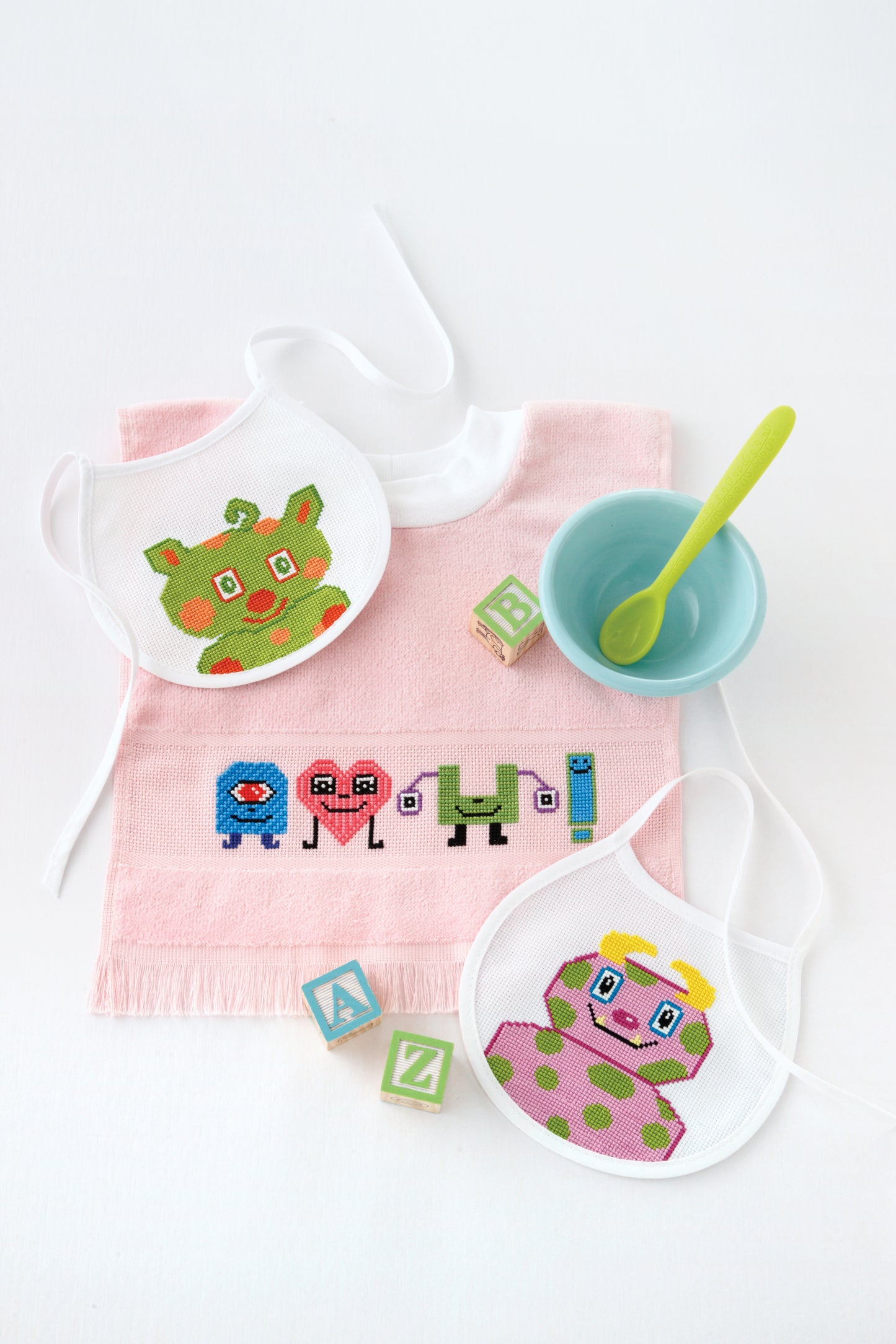 Leisure Arts' Lil' Monster Bibs Digital Download features playful monster cross stitch designs, shown with a pink cloth, pastel alphabet blocks, green spoon, and pale blue bowl on a white background.