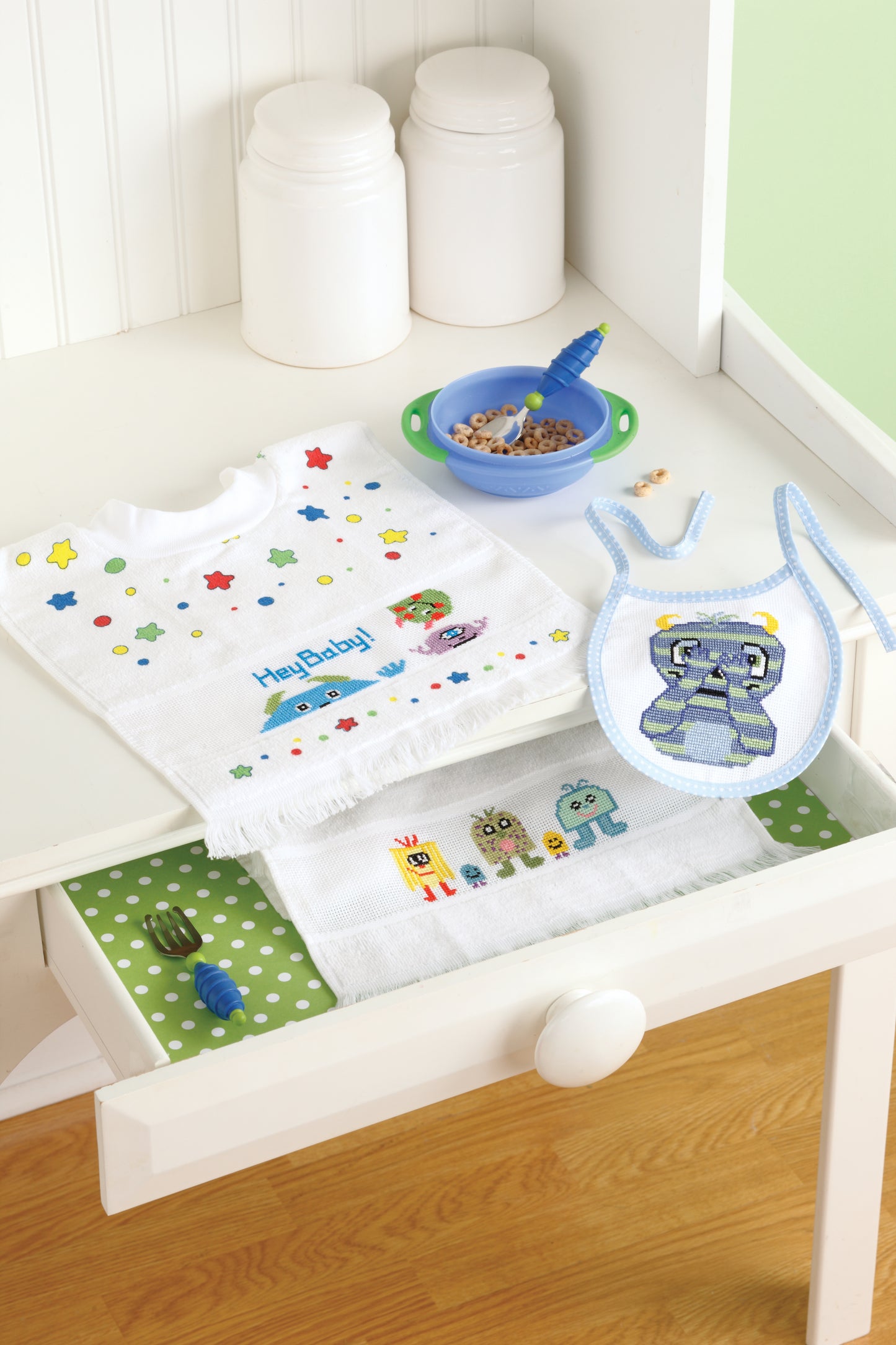 A white nursery drawer holds three Lil' Monster Bibs by Leisure Arts with cross-stitch designs, a green polka dot mat, fork, and blue spoon. On the counter are a baby bowl with cereal and two white containers.