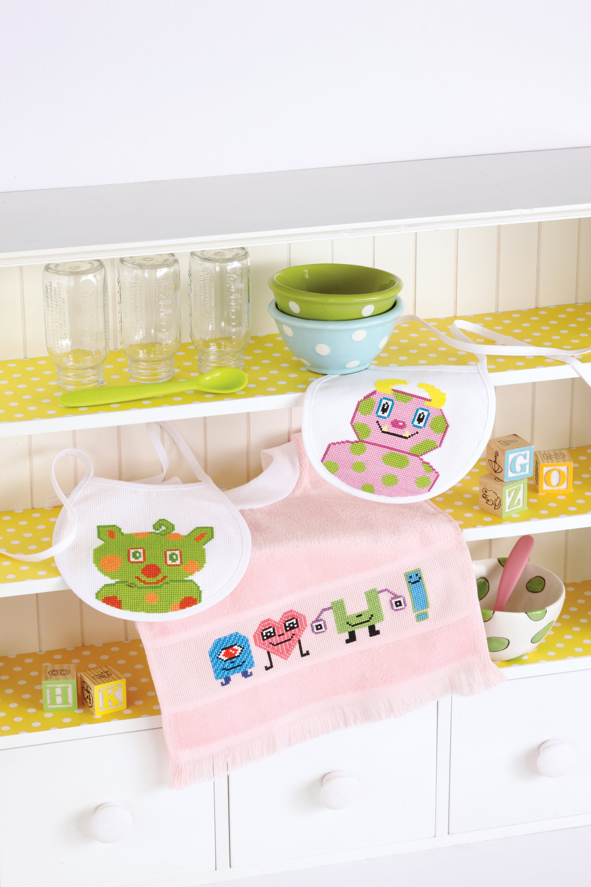 A white shelf with yellow polka-dot paper displays Leisure Arts' Lil' Monster Bibs Digital Download, featuring playful animal and letter cross stitch designs, along with glass jars, green bowls, a spoon, and alphabet blocks.