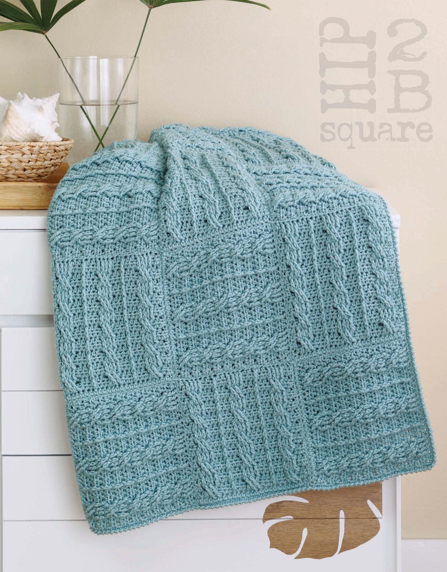 A Hip 2 B Square Throws 4 Home blanket by Leisure Arts, featuring a textured square pattern in blue medium weight yarn, is draped over white furniture with a basket and vase nearby. "2B HIP SQUARE" decorates the wall behind.