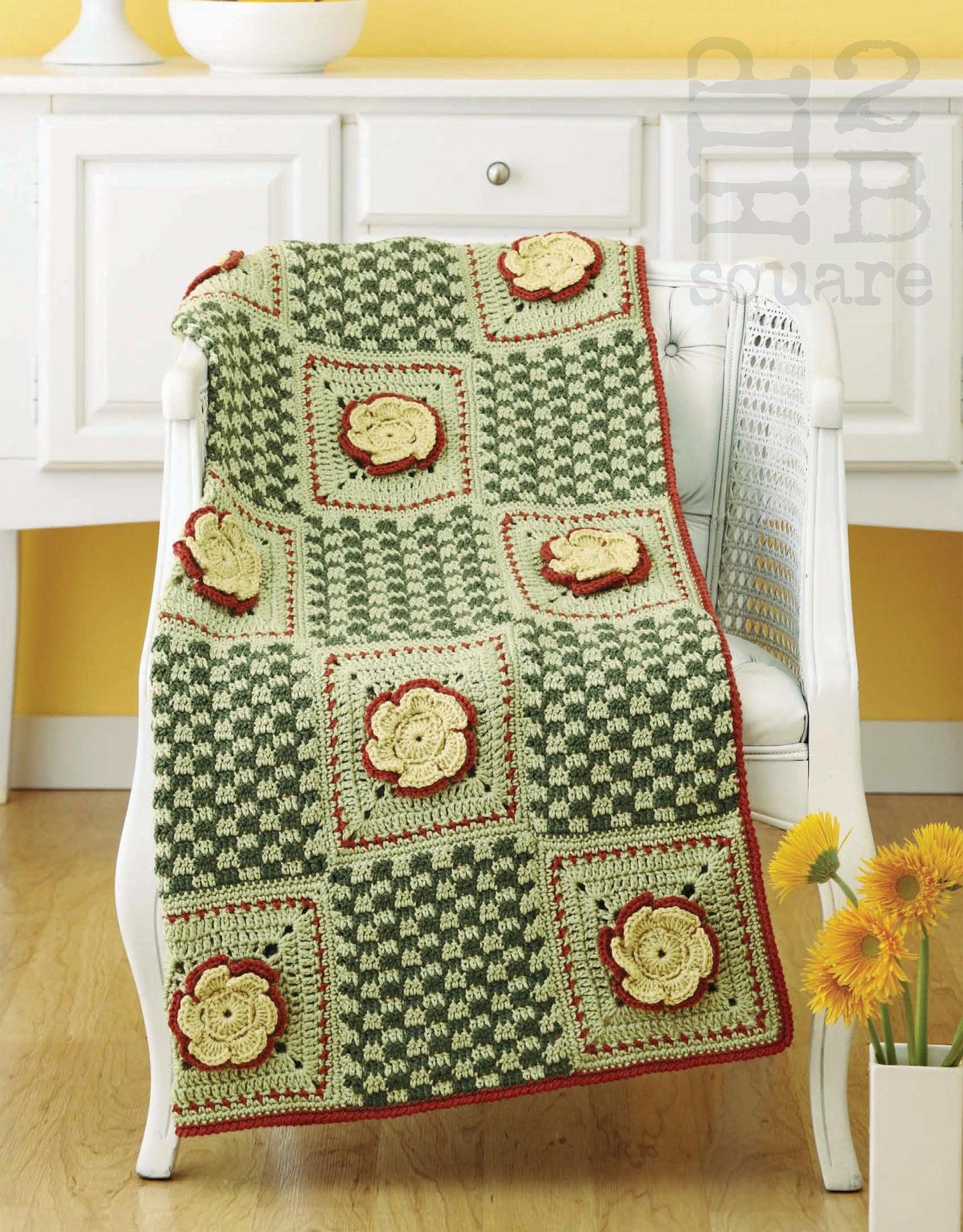 The Hip 2 B Square Throws 4 Home by Leisure Arts is a crocheted blanket with green and cream squares, yellow flower motifs outlined in red, shown draped over a white chair with a white cabinet and vase of yellow flowers in the background.