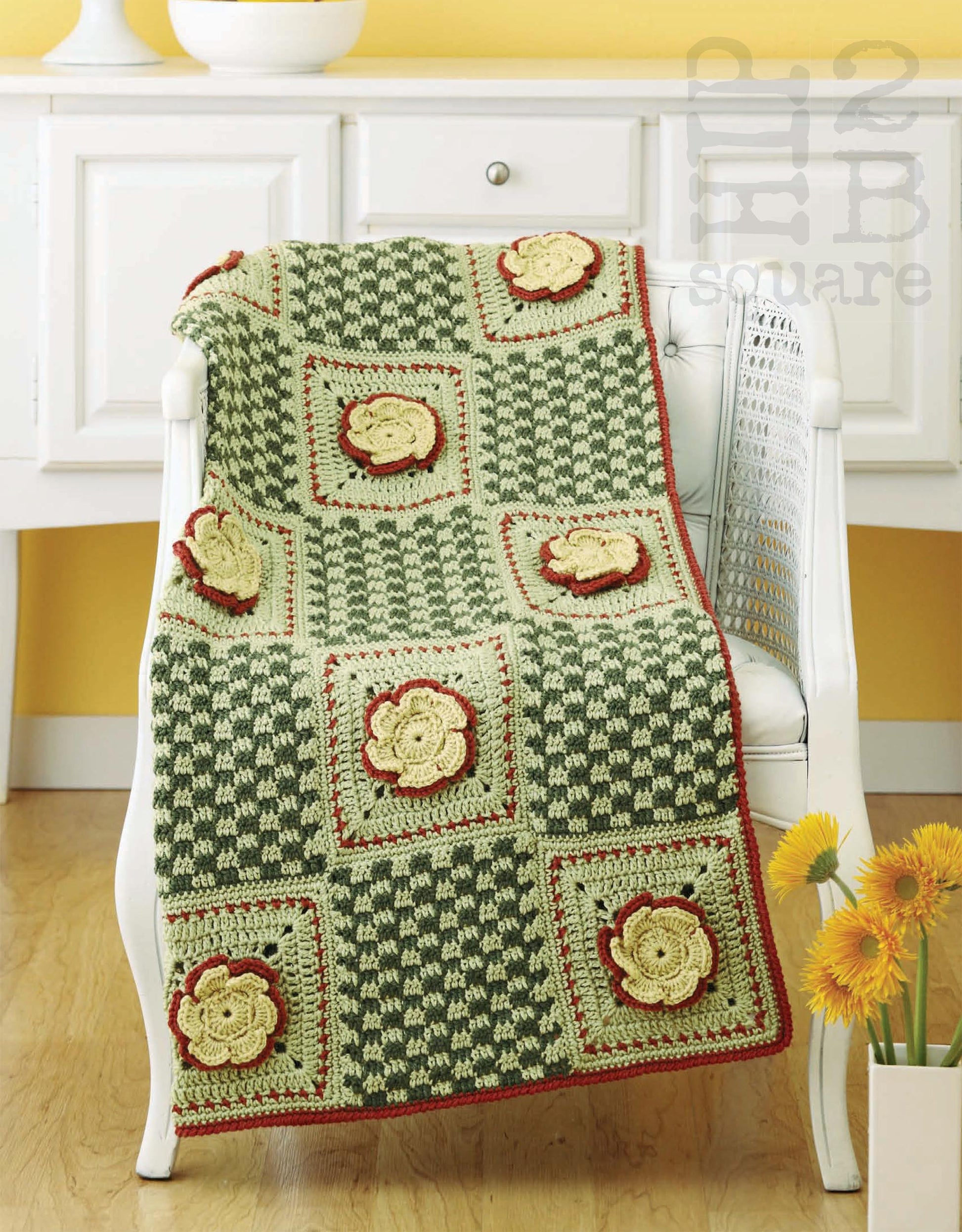 The Hip 2 B Square Throws 4 Home by Leisure Arts is a crocheted blanket with green and cream squares, yellow flower motifs outlined in red, shown draped over a white chair with a white cabinet and vase of yellow flowers in the background.