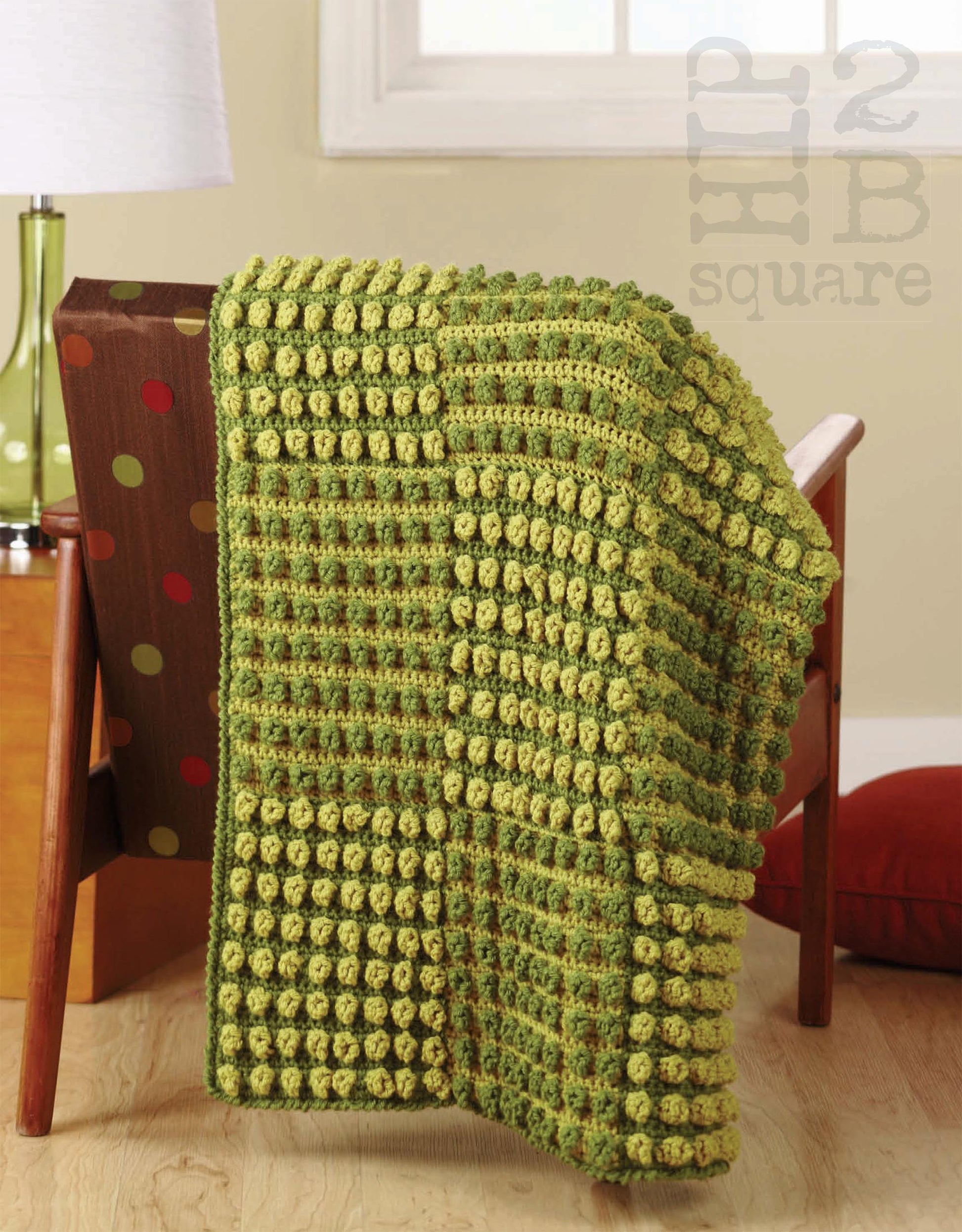 The Hip 2 B Square Throws 4 Home by Leisure Arts, a textured green and yellow crocheted blanket with raised bobbles and squares, is draped over a brown chair with colorful polka dots. A small lamp, red cushion, and window are in the background.