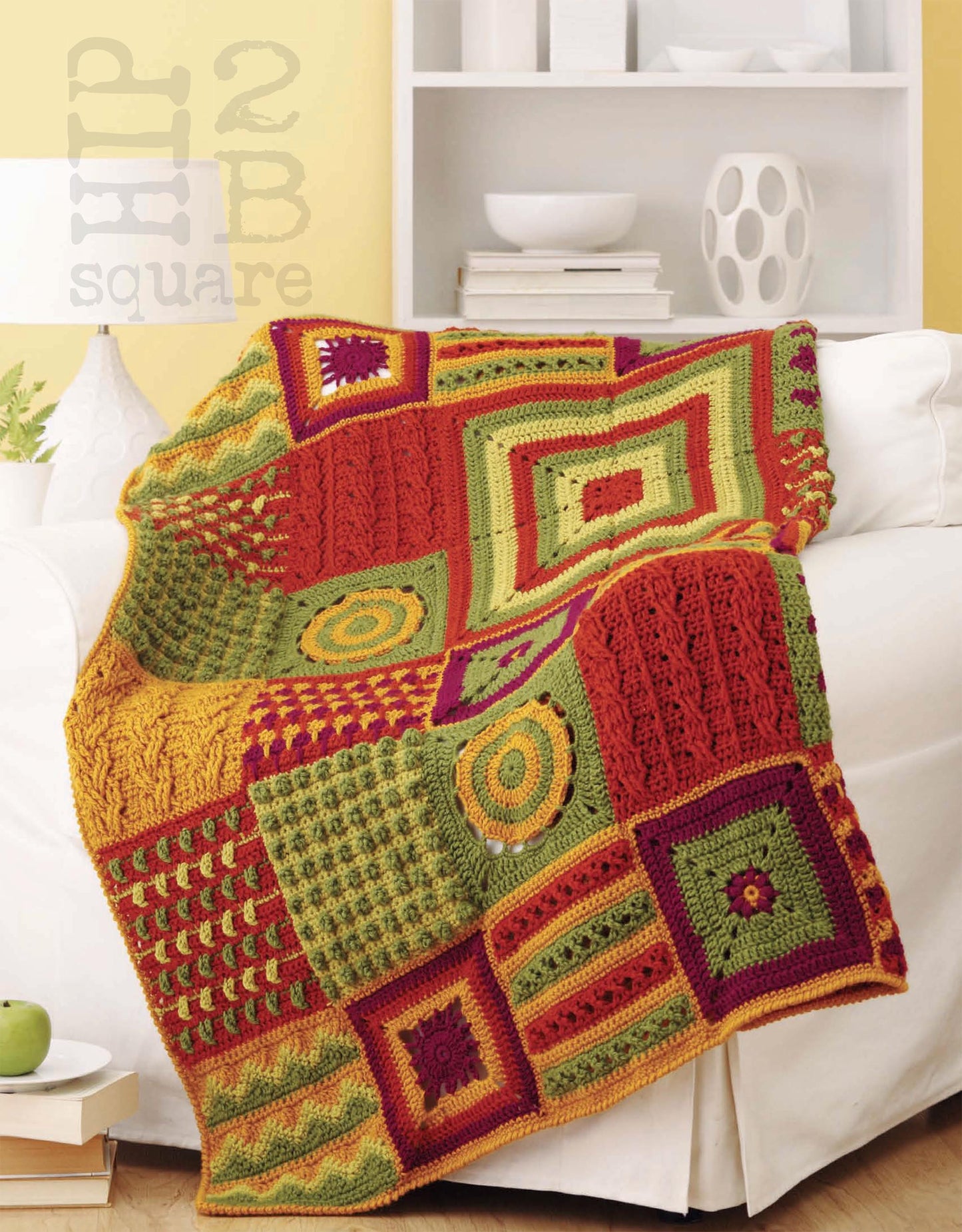 The Hip 2 B Square Throws 4 Home by Leisure Arts, featuring colorful red, green, yellow, and orange crochet squares in medium weight yarn, is draped over a white sofa in a bright room with yellow walls and modern decor.