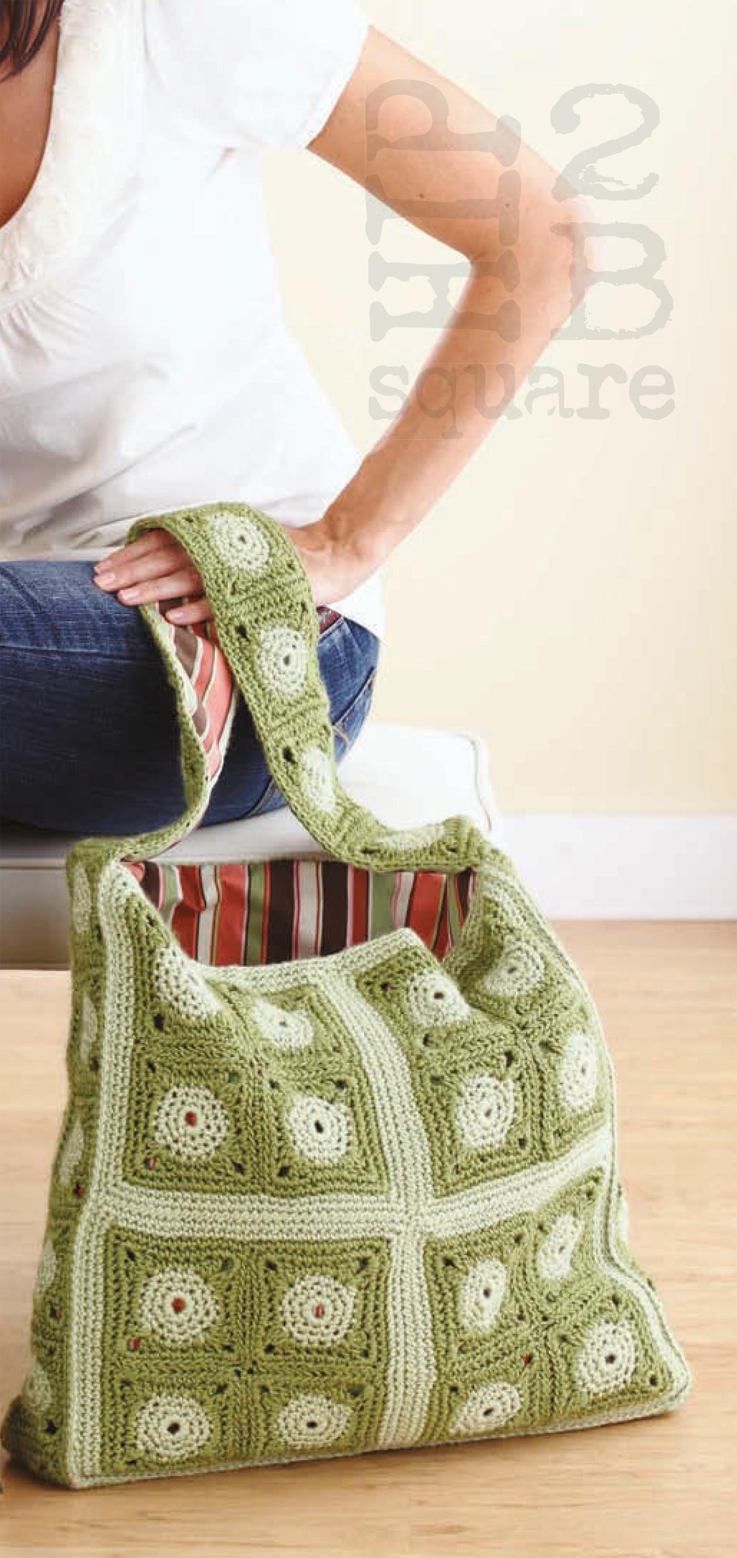 A person sits on the floor holding a Leisure Arts Hip 2 B Square Totes & Bags crochet bag with green and white square motifs, a striped lining, and a wide handle.