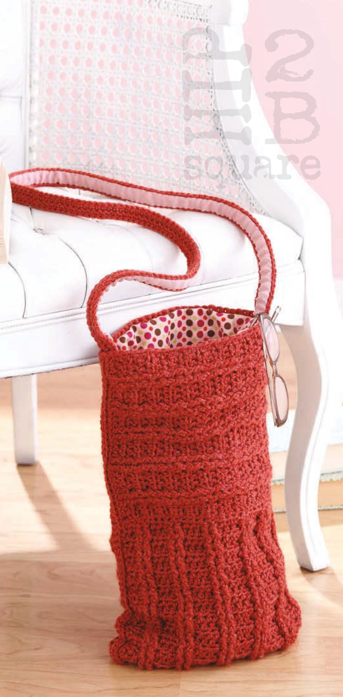 The Hip 2 B Square Totes & Bags Digital Download by Leisure Arts features a red, textured crochet shoulder bag with detailed motifs and a patterned lining, displayed on a cane-back chair—a stylish idea for your next crochet project.