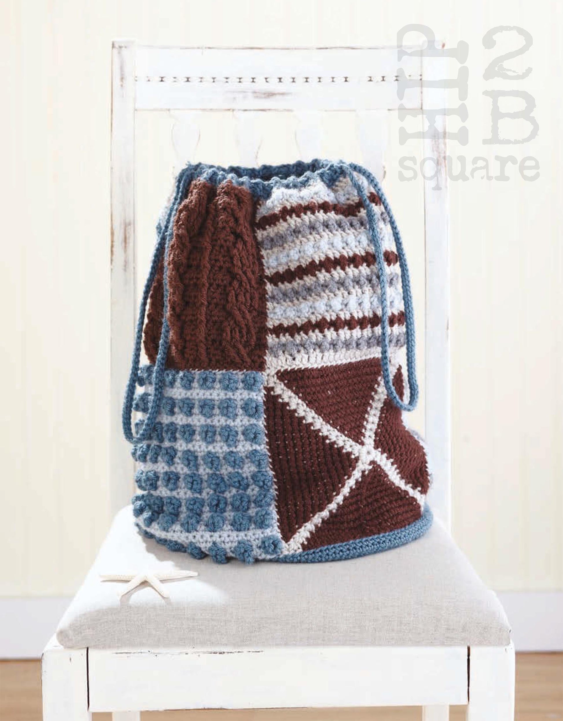 The Hip 2 B Square Totes & Bags Digital Download from Leisure Arts is displayed on a white wooden chair, showcasing its brown, blue, and white patchwork crochet motifs with textured squares, stripes, and a blue cord. A small white starfish rests nearby.