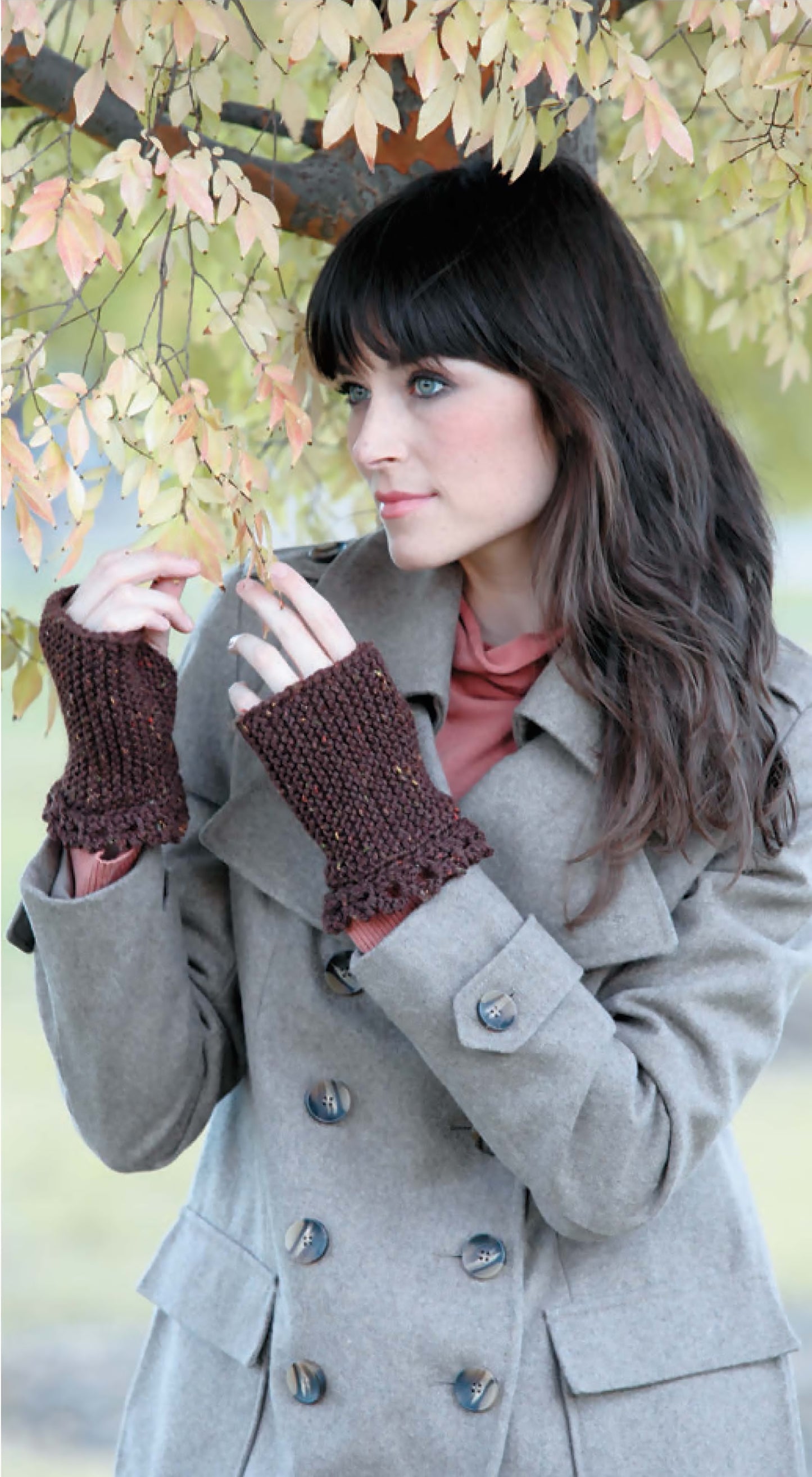 Wearing a light gray coat and brown mittens made with Leisure Arts’ "I Can't Believe I'm Knitting Mittens," perfect for beginners, a woman stands under a tree with yellow leaves, gently touching a branch as she gazes thoughtfully to the side.