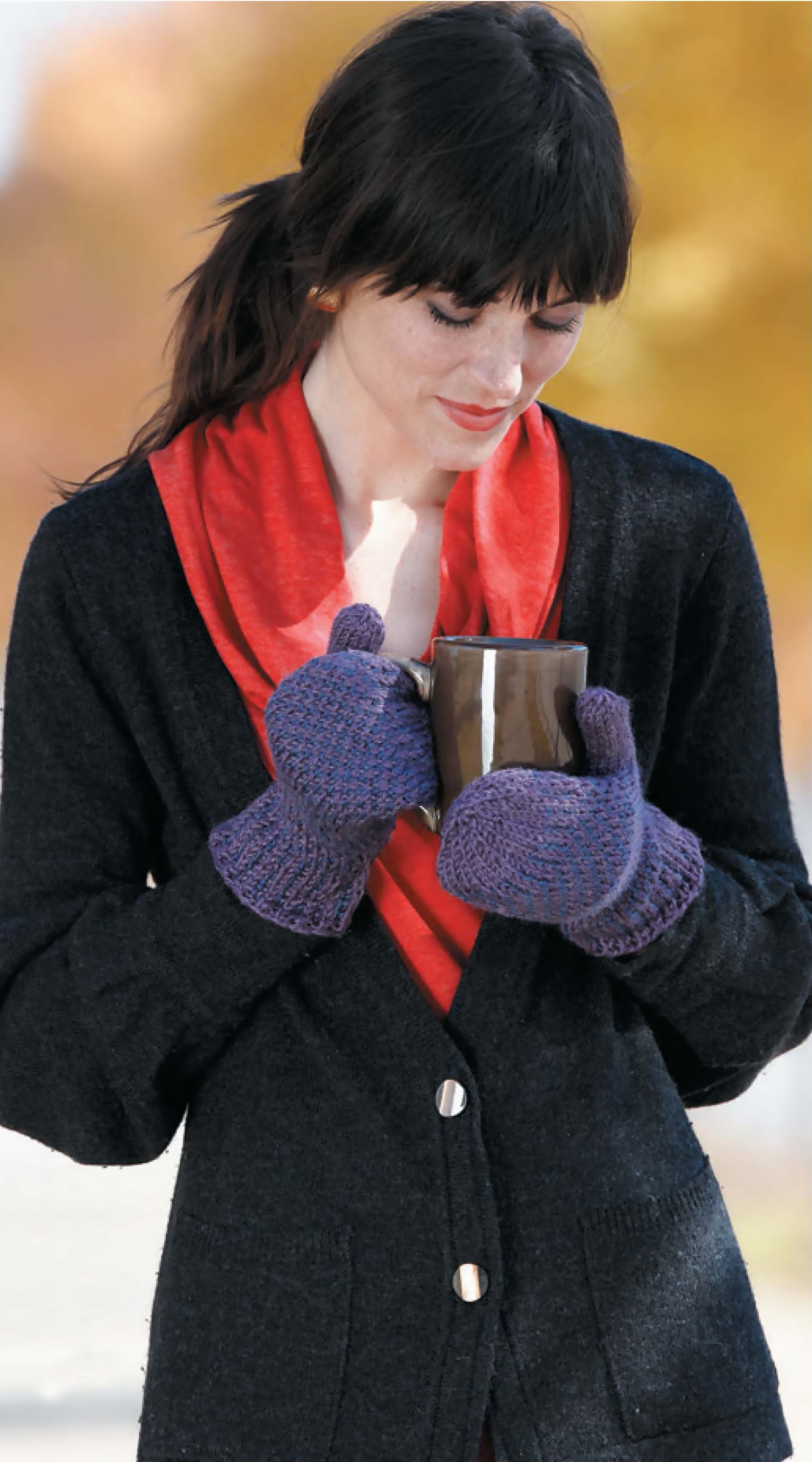 A woman in a red scarf, black cardigan, and purple mittens made from Leisure Arts' "I Can't Believe I'm Knitting Mittens" smiles down at her mug against an autumn backdrop of yellow and orange trees.