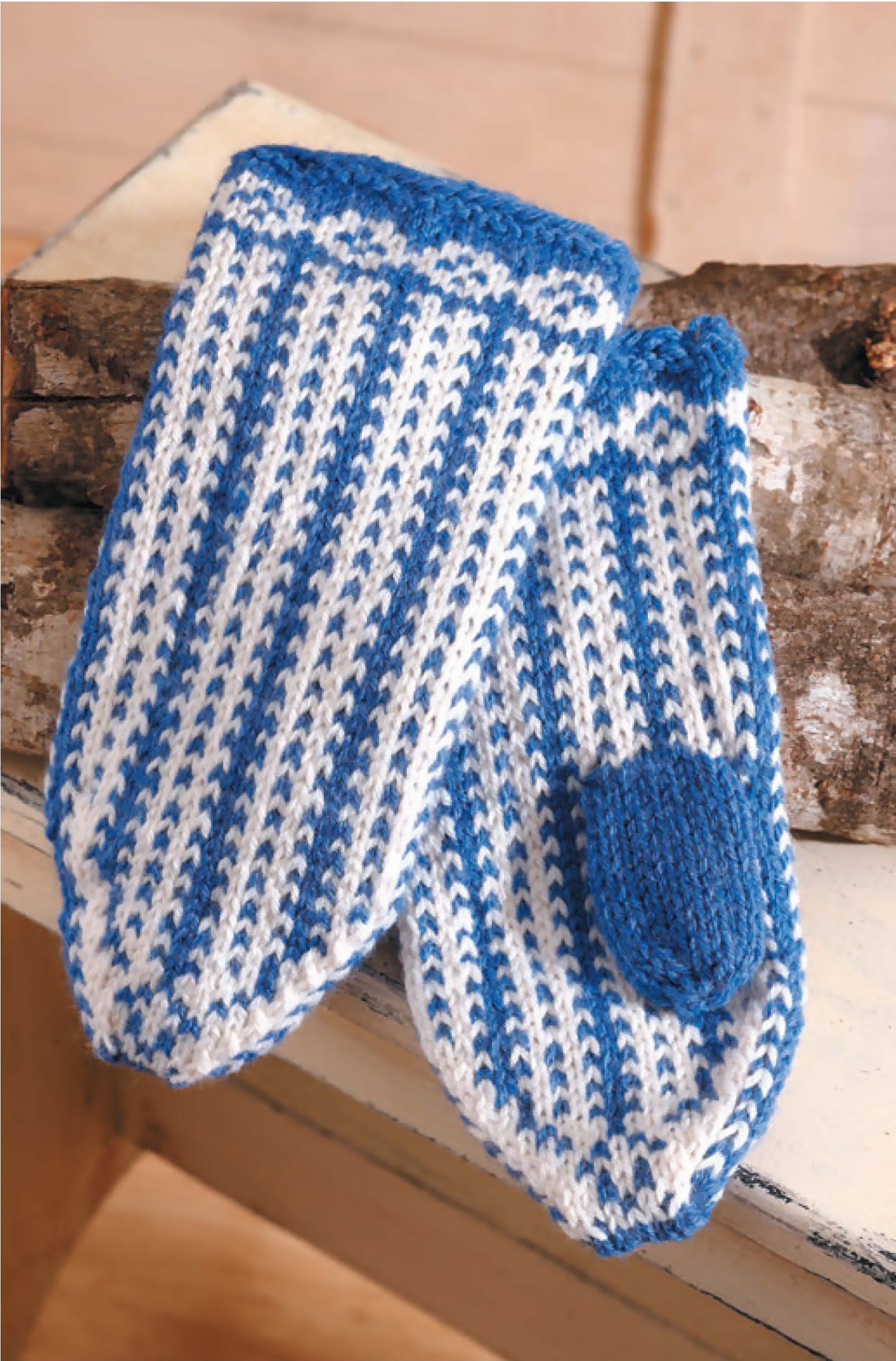 A pair of blue and white striped mittens with solid blue cuffs and thumbs rests on a birch log, featured in Leisure Arts' "I Can't Believe I'm Knitting Mittens"—perfect for those exploring basic mitten knitting patterns on a white surface.