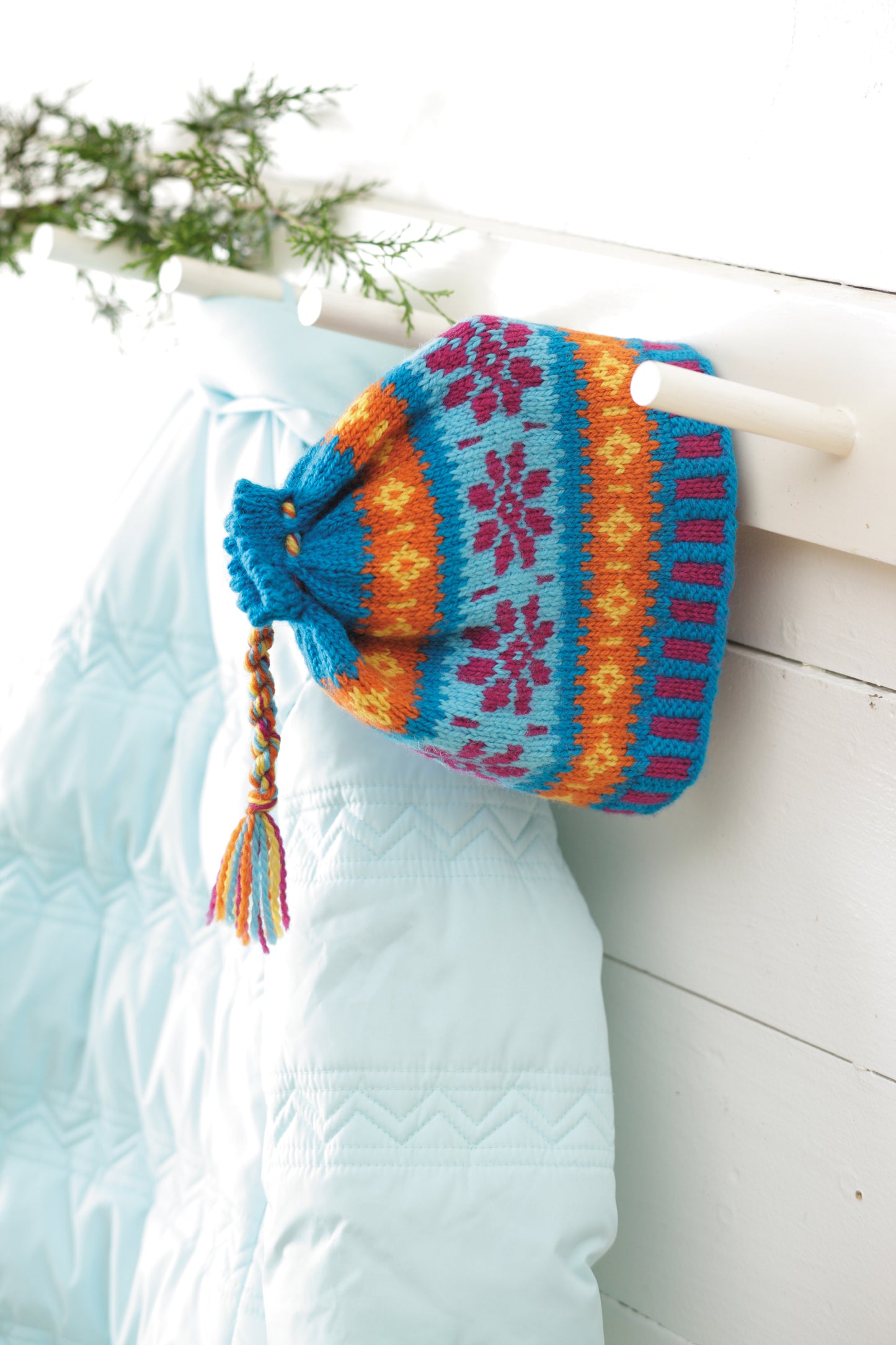 The "I Can't Believe I'm Fair Isle Knitting Digital Download" by Leisure Arts features vibrant blue, orange, and red knit hat patterns—ideal for winter projects—displayed on a white peg beside a light blue quilted jacket and green pine branch.