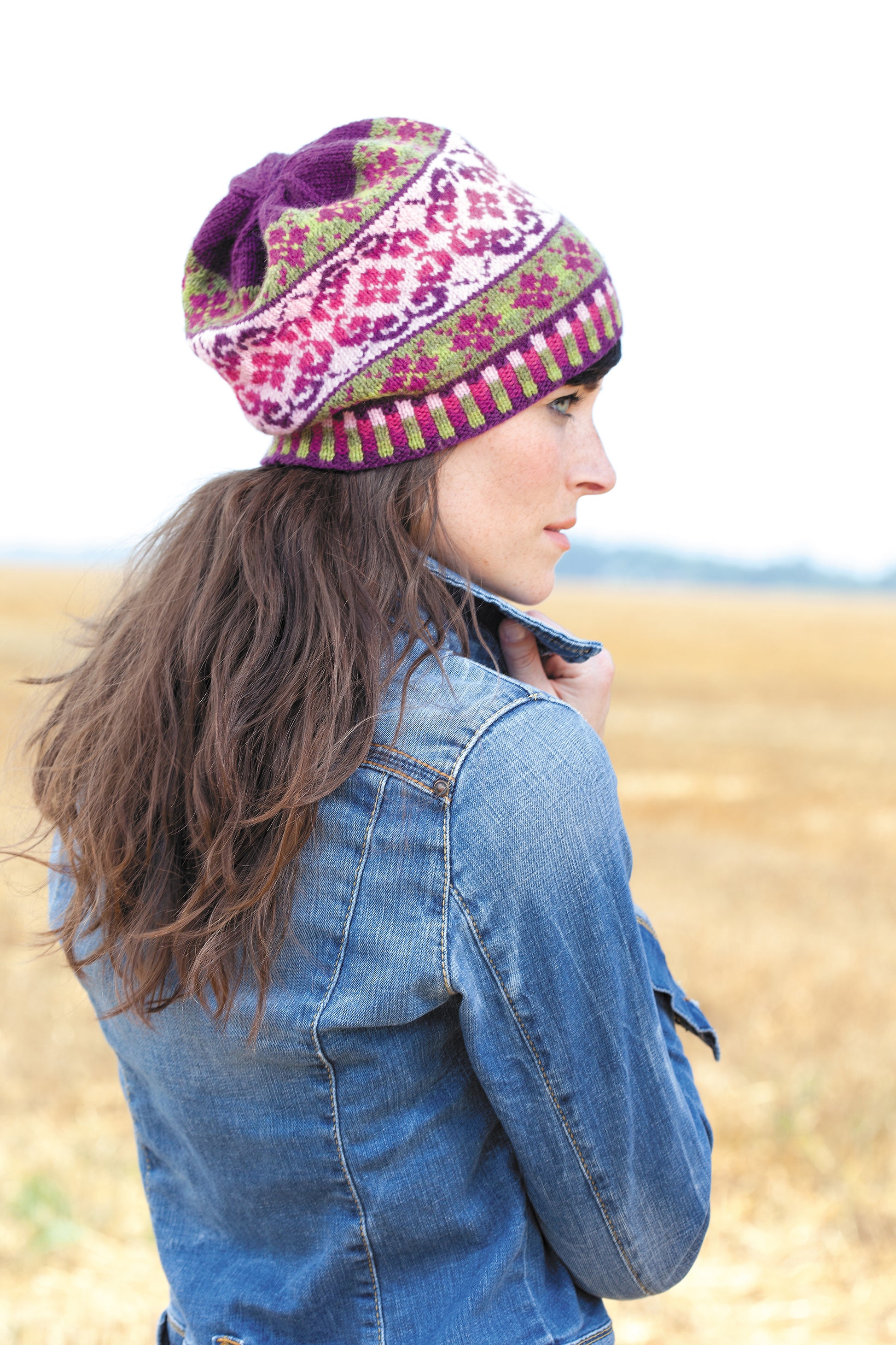 A woman with long brown hair wears a colorful knit hat inspired by the "I Can't Believe I'm Fair Isle Knitting" digital download by Leisure Arts, paired with a denim jacket, standing outdoors among dry grass under an overcast sky.