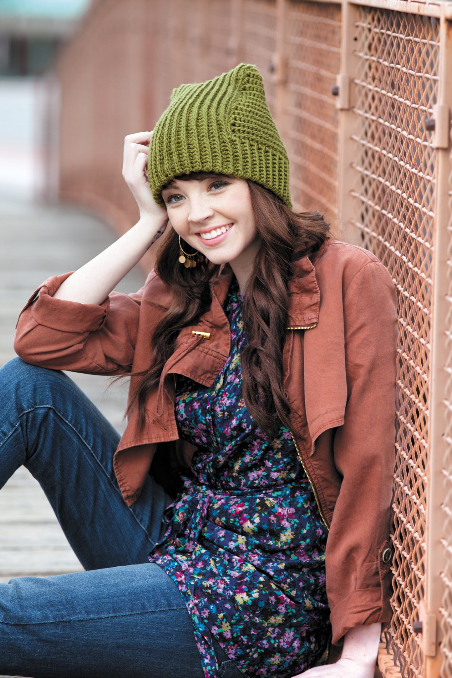 A smiling woman with long brown hair wears a green beanie inspired by styles from Heady Affairs—7 Impetuous Hats to Crochet by Leisure Arts, along with a brown jacket, floral dress, and jeans as she sits on a wooden bridge by a metal fence.