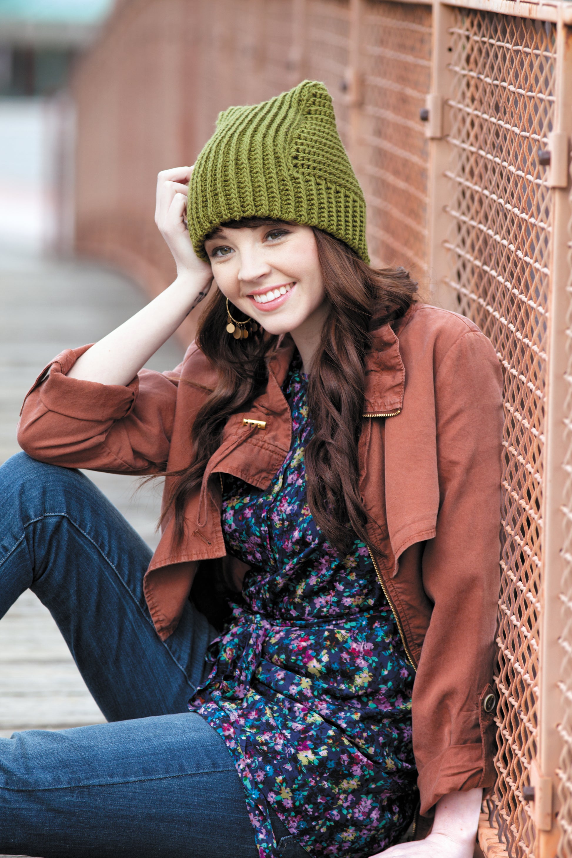 A smiling woman with long brown hair wears a green beanie inspired by styles from Heady Affairs—7 Impetuous Hats to Crochet by Leisure Arts, along with a brown jacket, floral dress, and jeans as she sits on a wooden bridge by a metal fence.