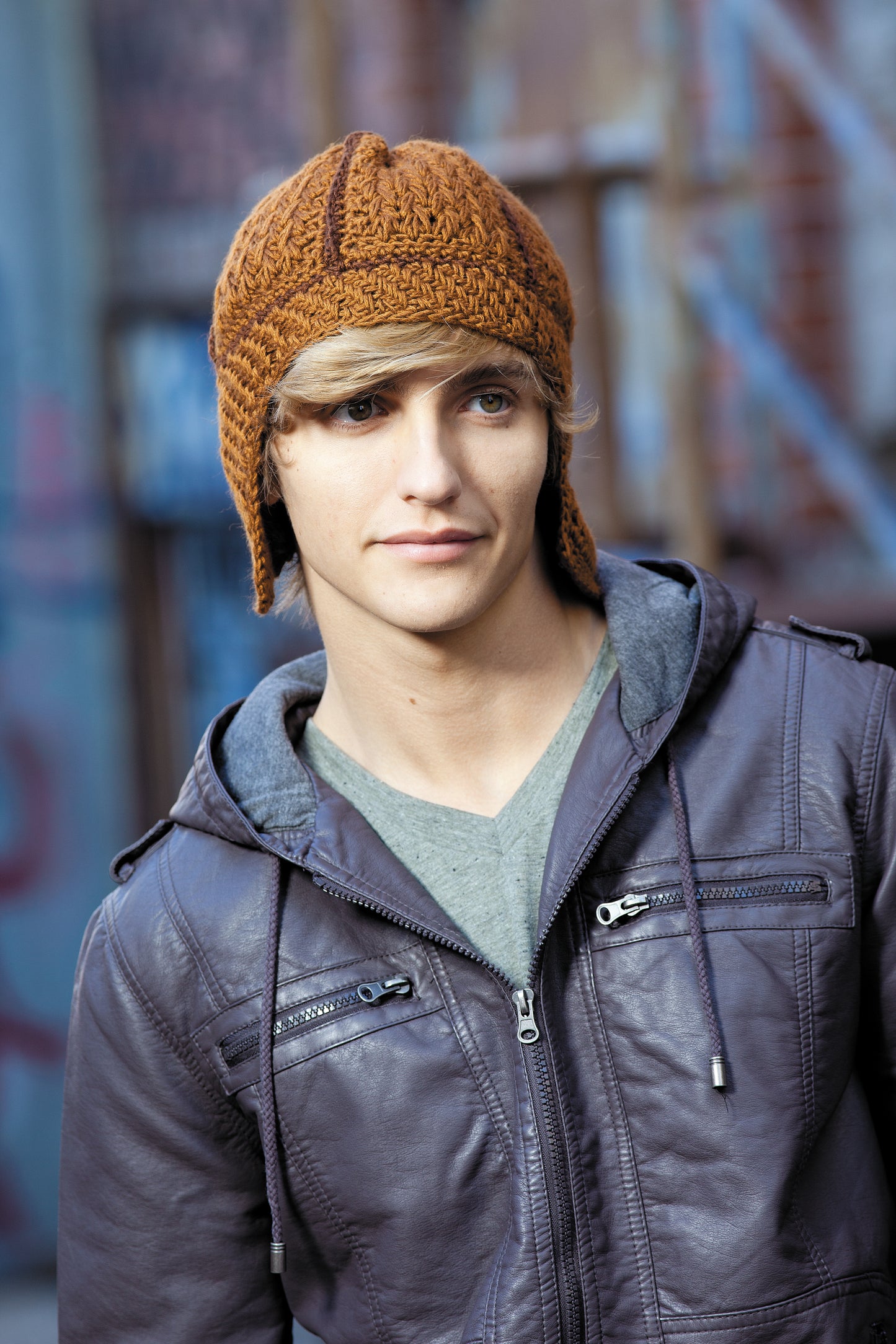 A young person stands in front of urban graffiti, wearing a brown crochet earflap hat made with worsted yarn from "Heady Affairs—7 Impetuous Hats to Crochet" by Leisure Arts, paired with a gray T-shirt and brown leather jacket.