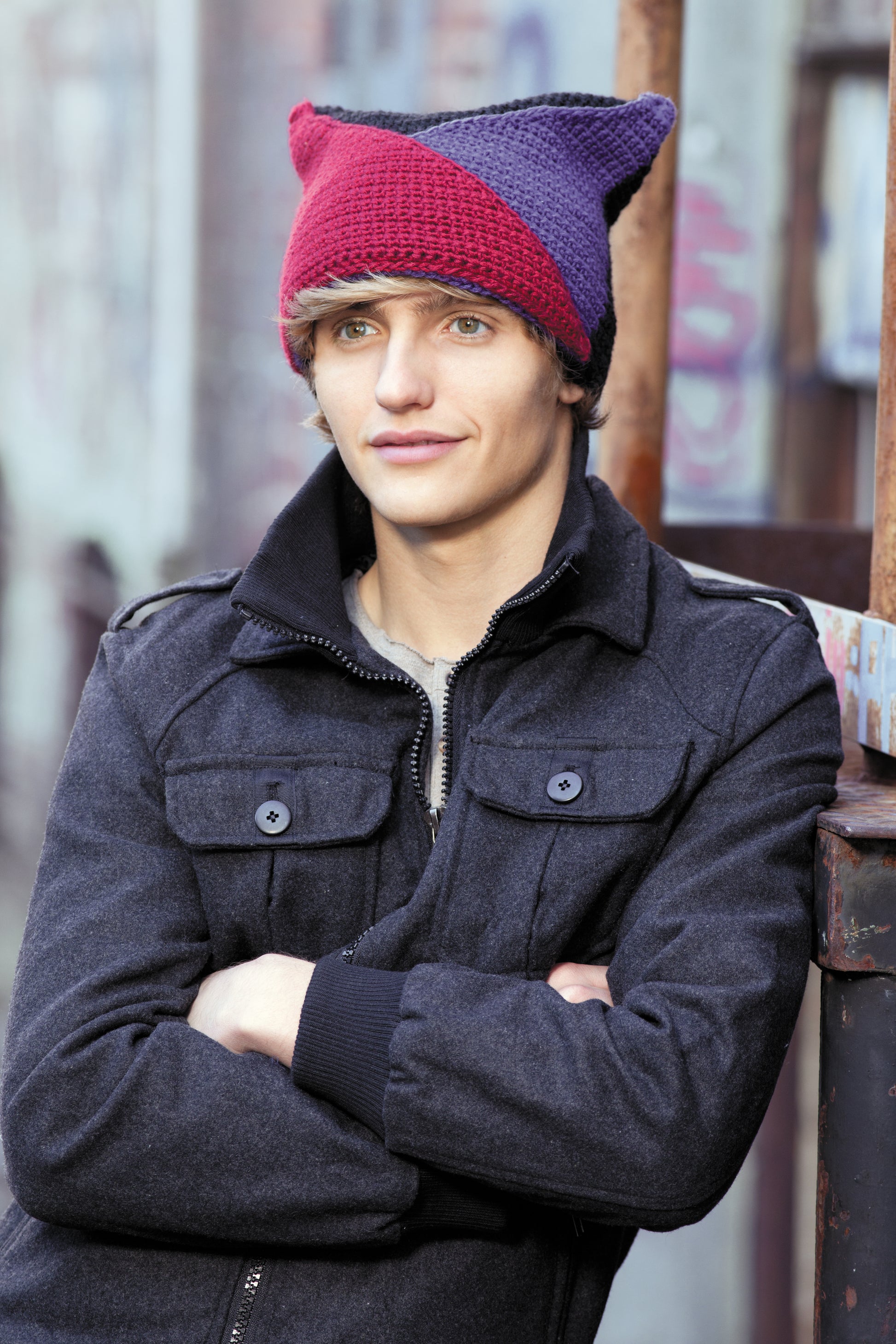 Wearing a two-toned red and purple crochet hat from "Heady Affairs—7 Impetuous Hats to Crochet" by Leisure Arts, a person in a dark jacket stands outdoors with arms crossed, set against a blurred graffiti wall.