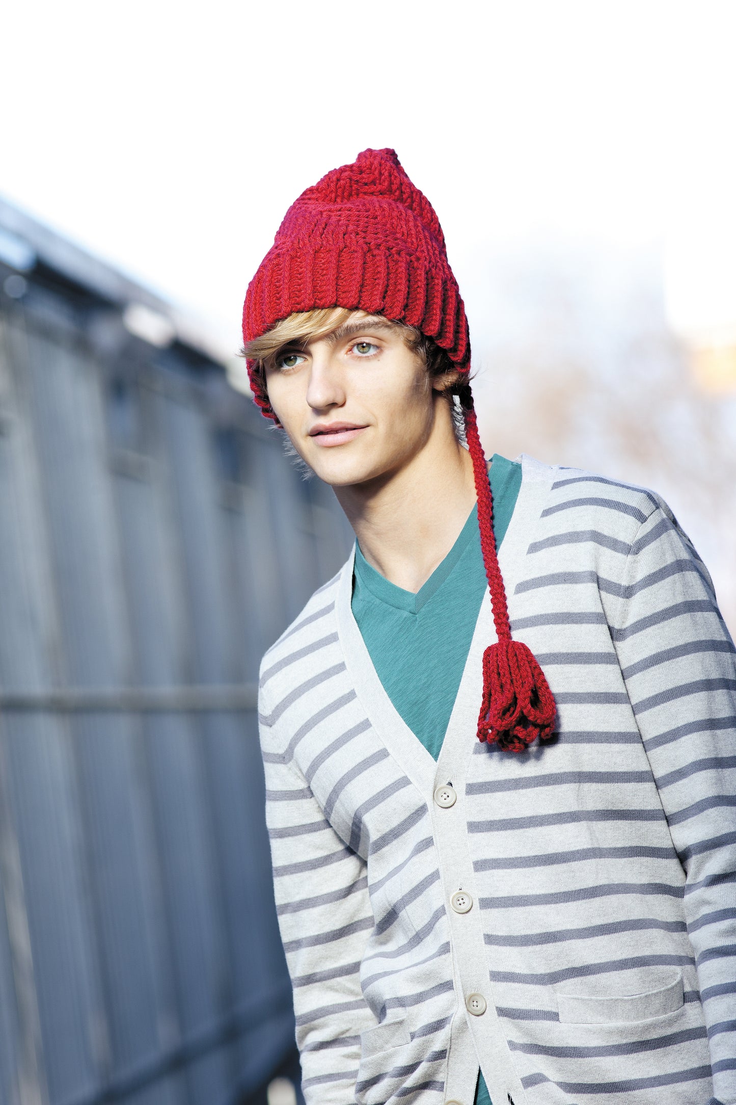 A young person in a red crochet hat from "Heady Affairs—7 Impetuous Hats to Crochet" by Leisure Arts, a teal shirt, and a striped cardigan stands outdoors by a metal fence with a softly blurred background.