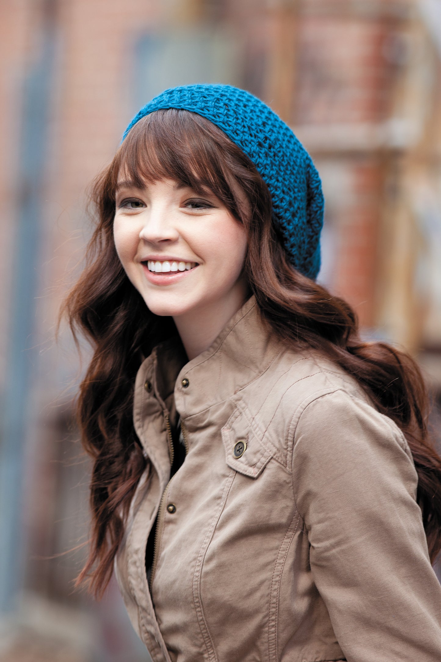 A young woman with long brown hair wears a blue crocheted beanie made from Heady Affairs—7 Impetuous Hats to Crochet by Leisure Arts, smiling outdoors in a tan jacket against a blurred background.