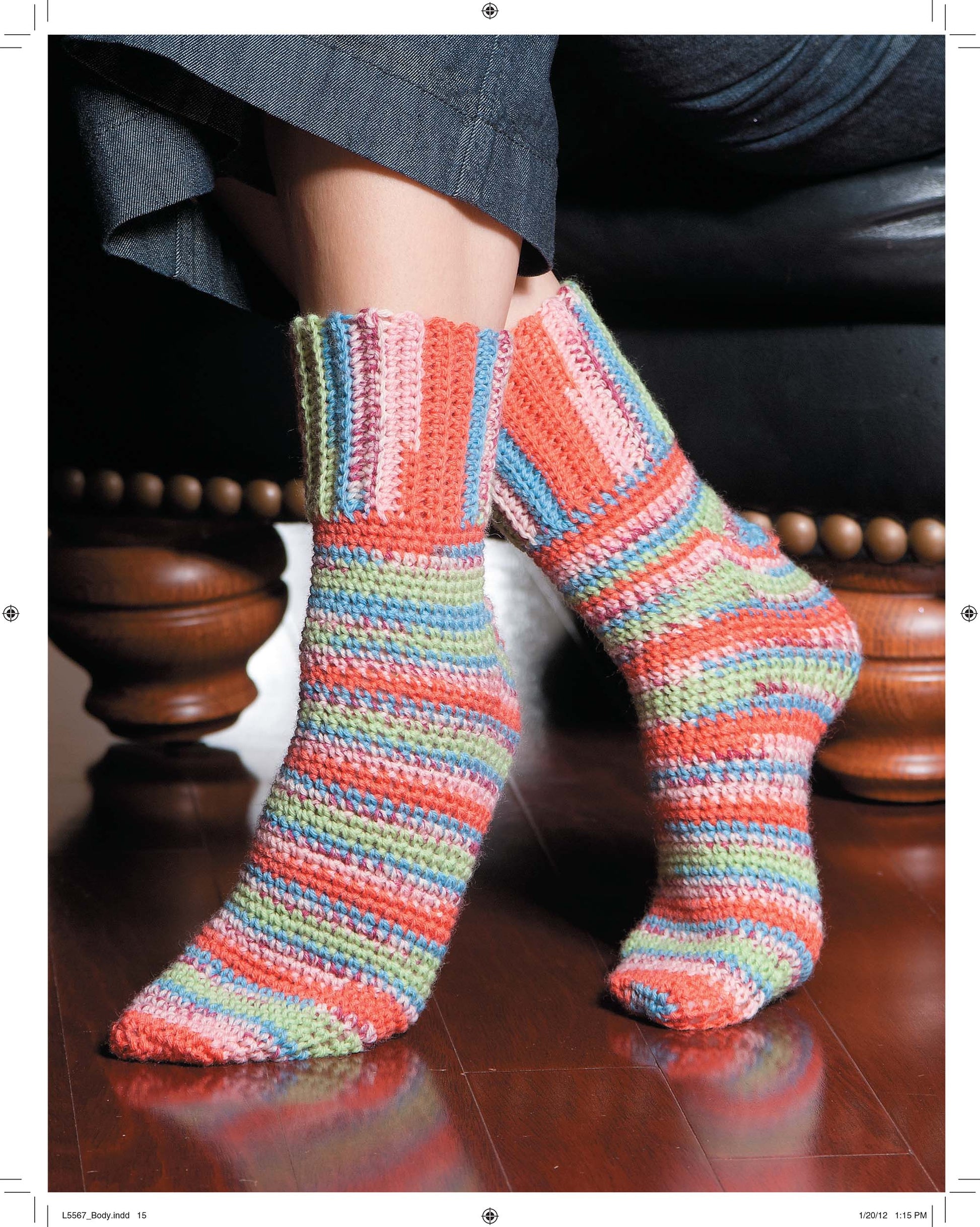 Someone wearing Leisure Arts’ “Socks for the Family”—colorful, hand-crocheted socks in blue, pink, green, orange, and red—sits on a dark leather chair with wooden legs.