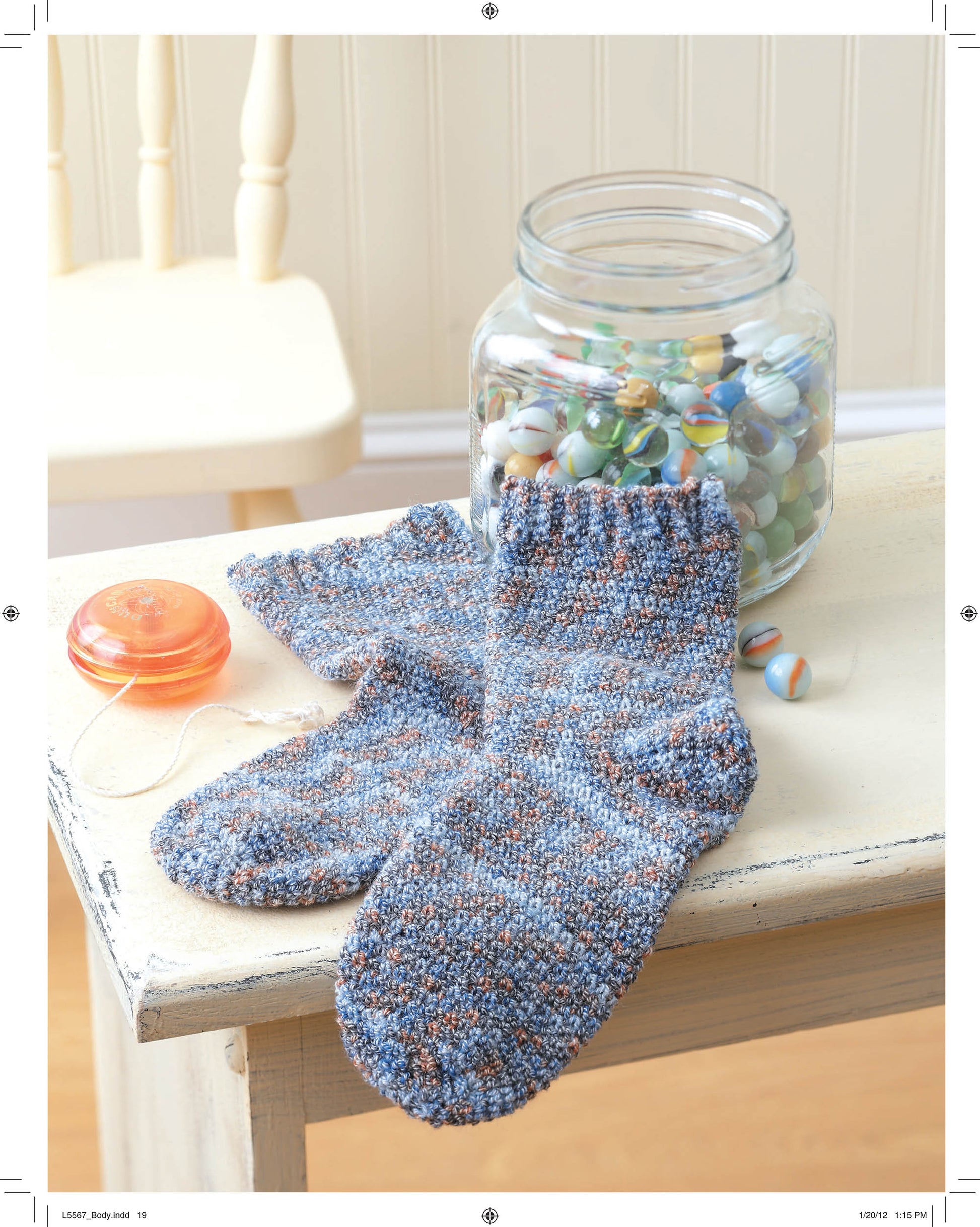 A pair of knitted socks from Leisure Arts’ "Socks for the Family" rests on a wooden table beside a jar of marbles and an orange yo-yo, inspiring you to create your own crochet patterns. A white chair is visible in the background.